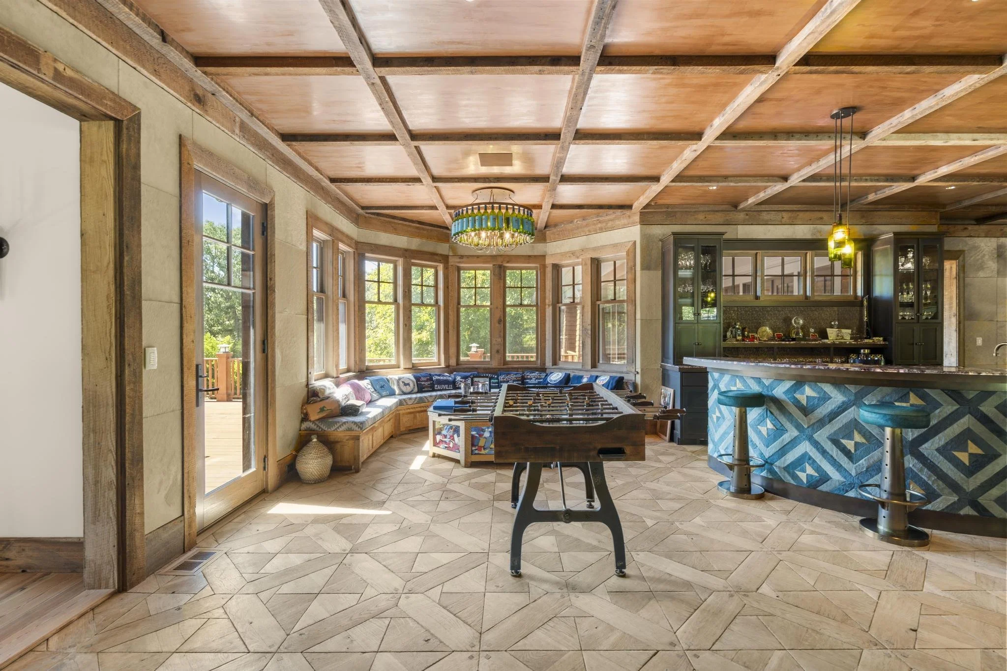 Living room with coffered wooden ceiling, large windows, a built-in window seat with pillows, a foosball table, and a view of an outdoor deck.