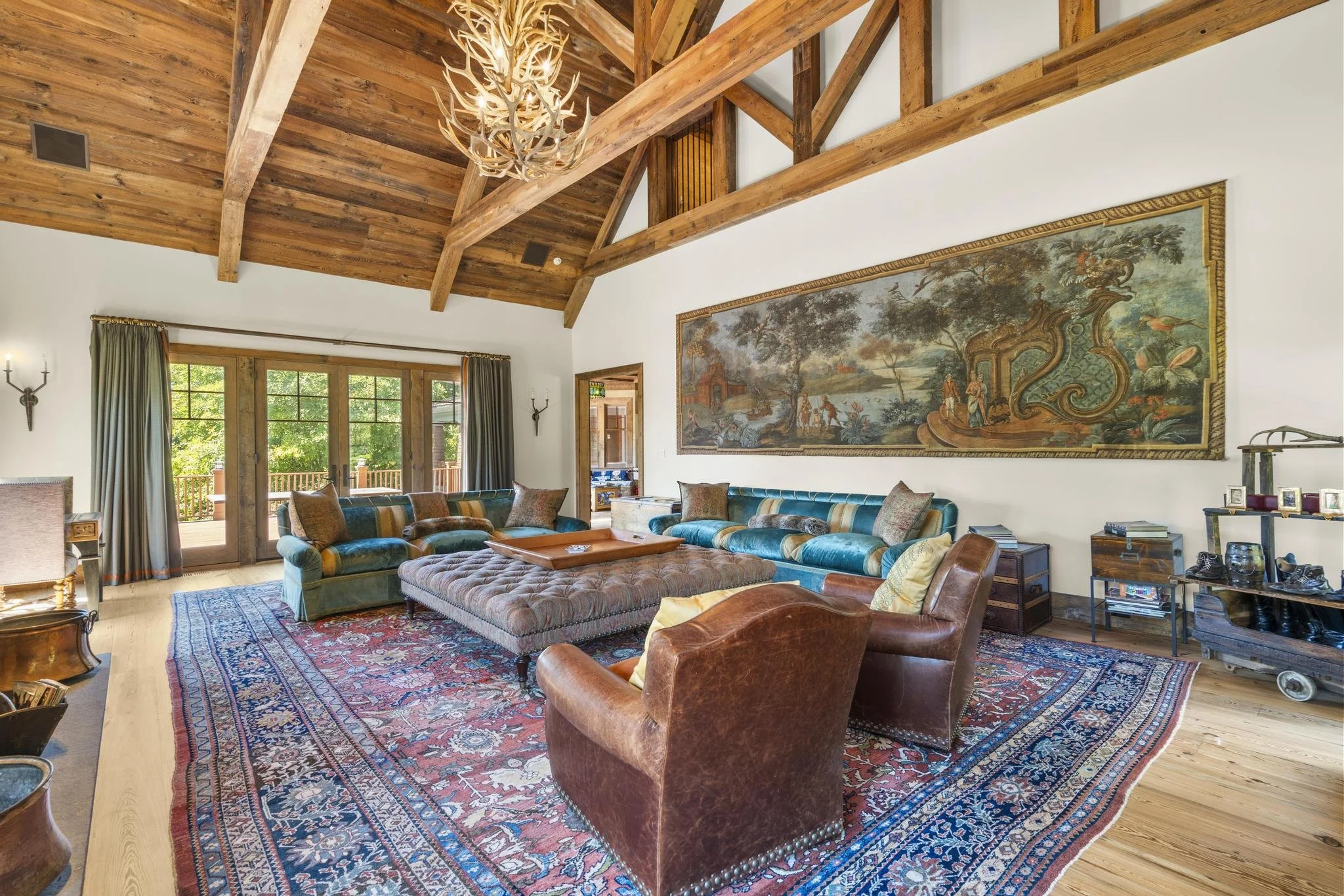 Living room with wooden vaulted ceiling, chandelier, large framed tapestry on wall, multiple couches, and sliding glass doors leading to outdoor deck.