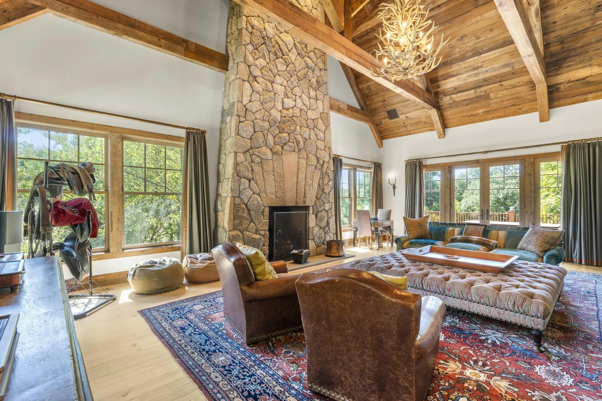 Living room with a large stone fireplace, wooden vaulted ceiling, and big windows showing trees outside. There are assorted chairs, a couch, and a colorful rug.