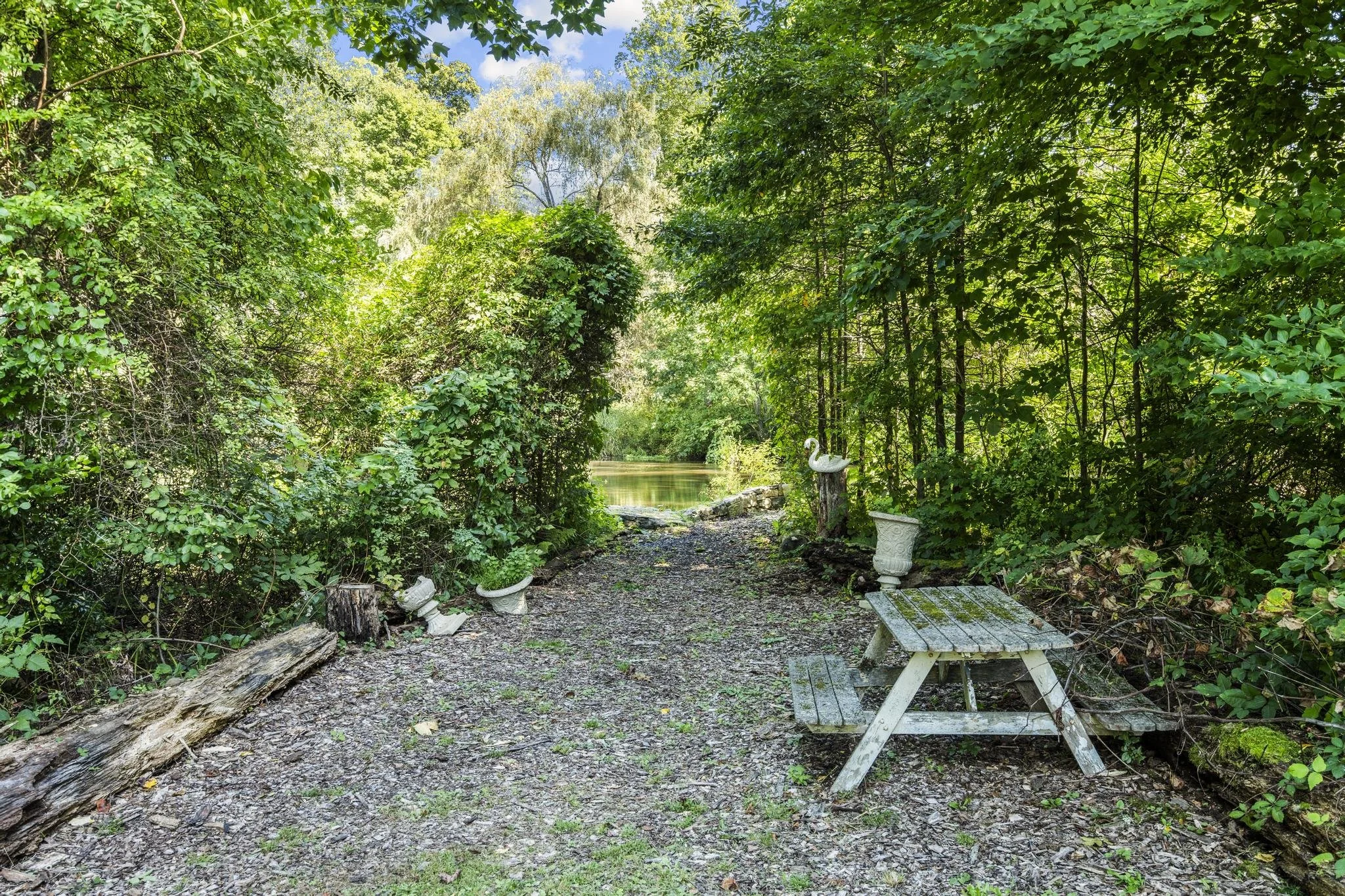 A lush green forest scene with a small stream, trees, and foliage. There are garden ornaments and a bench along the pathway.