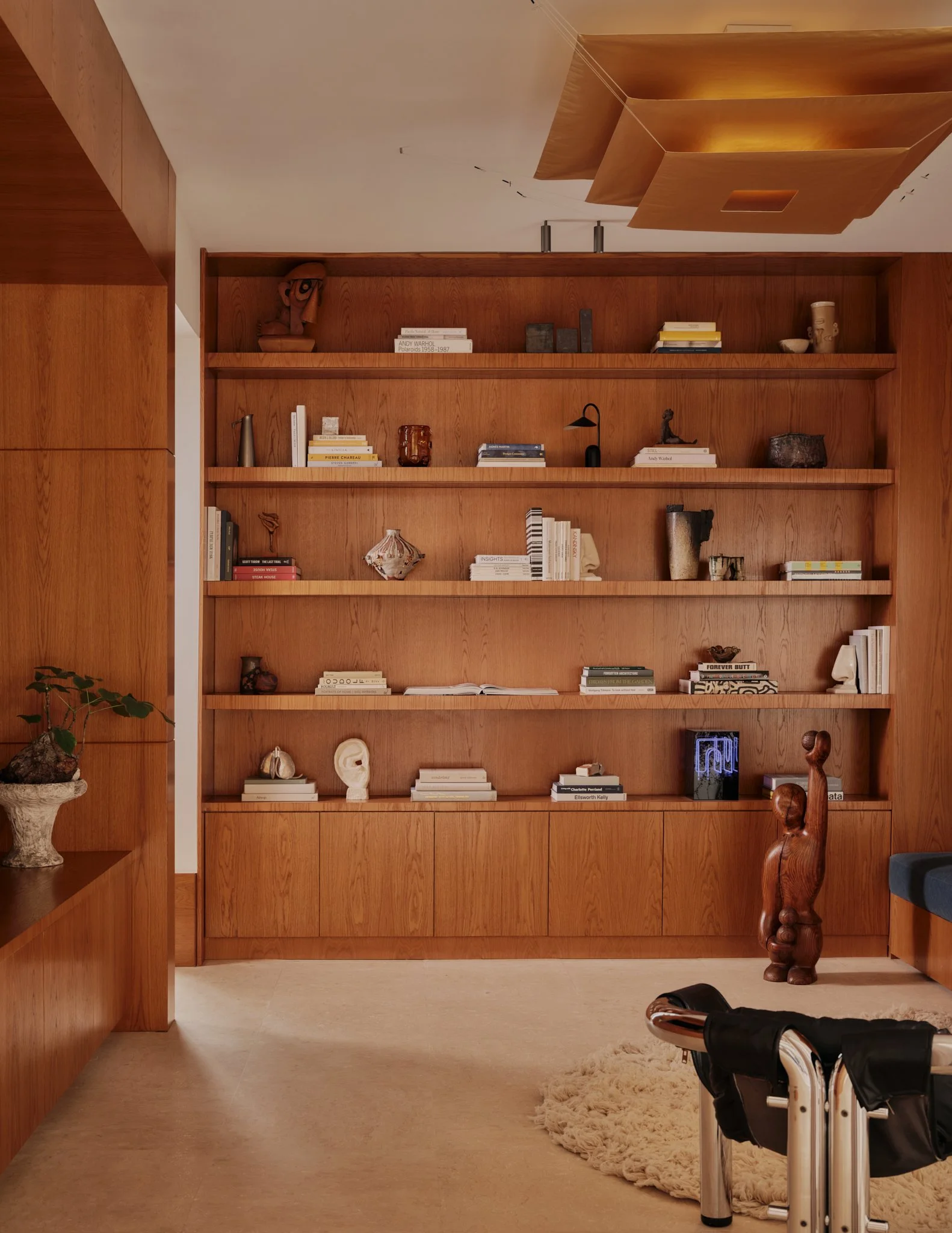 A wooden bookshelf with art books and decorative objects, a wooden sculpture of a stylized figure, a cozy rug, and a small table with a plant in a pot.