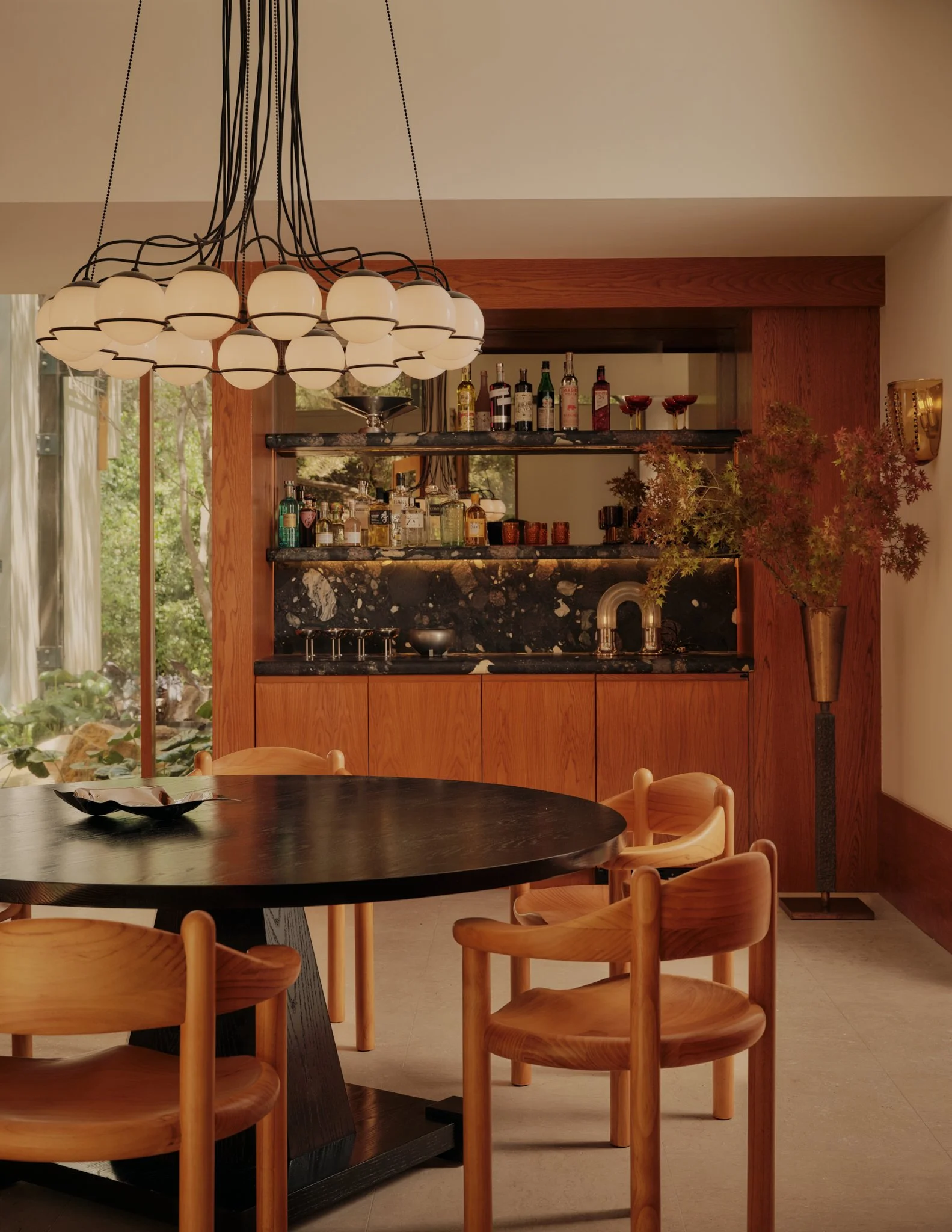 A modern kitchen with a black marble countertop, wooden cabinets, and a circular wooden dining table with matching chairs. A black chandelier with multiple white globes hangs above the table. The background features a bar area with various bottles of alcohol and glassware, and a large window with view of greenery outside.