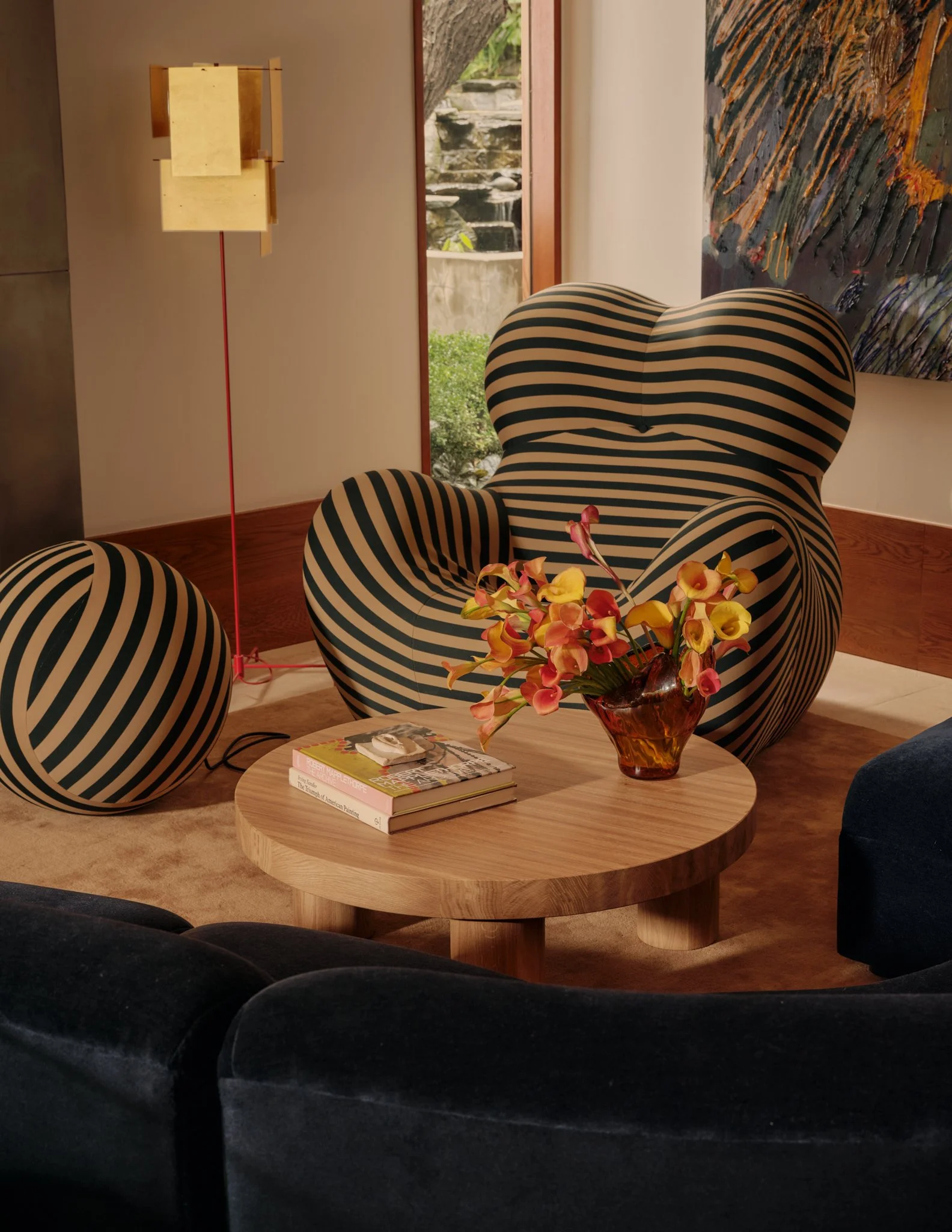 A living room setting with a black and white striped armchair, a round wooden coffee table with books and a flower arrangement, a beige floor lamp, a window with a view of a garden, and a colorful abstract painting on the wall.