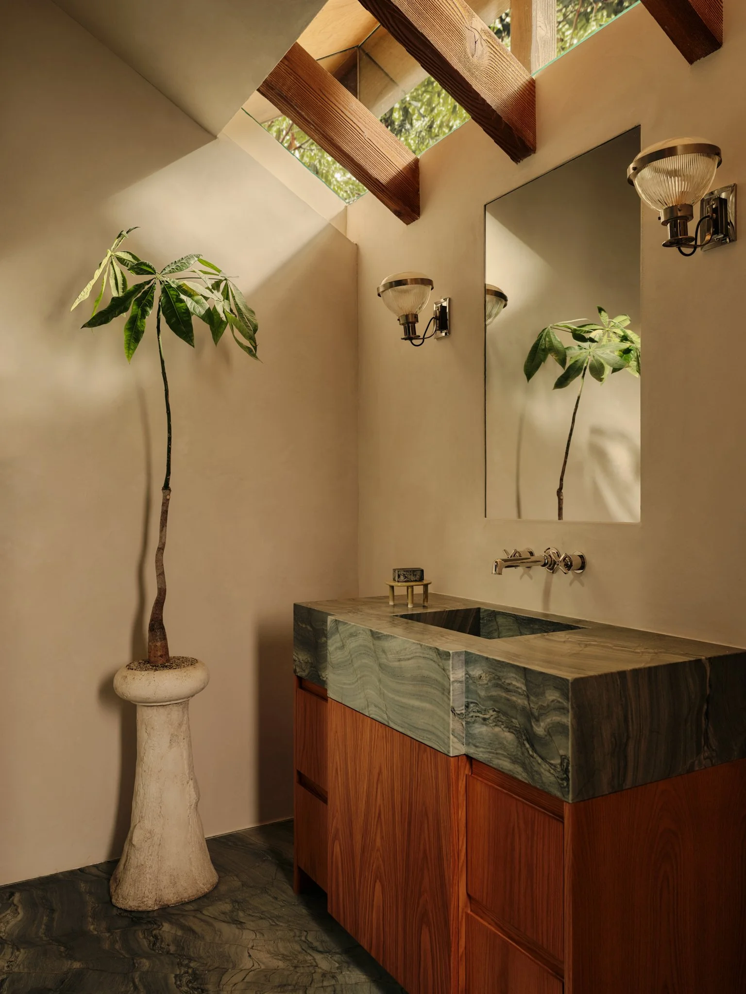 Interior view of a bathroom with a marble sink, a large mirror, wall-mounted light fixtures, a potted plant on a pedestal, and a ceiling skylight with wooden beams and greenery visible outside.