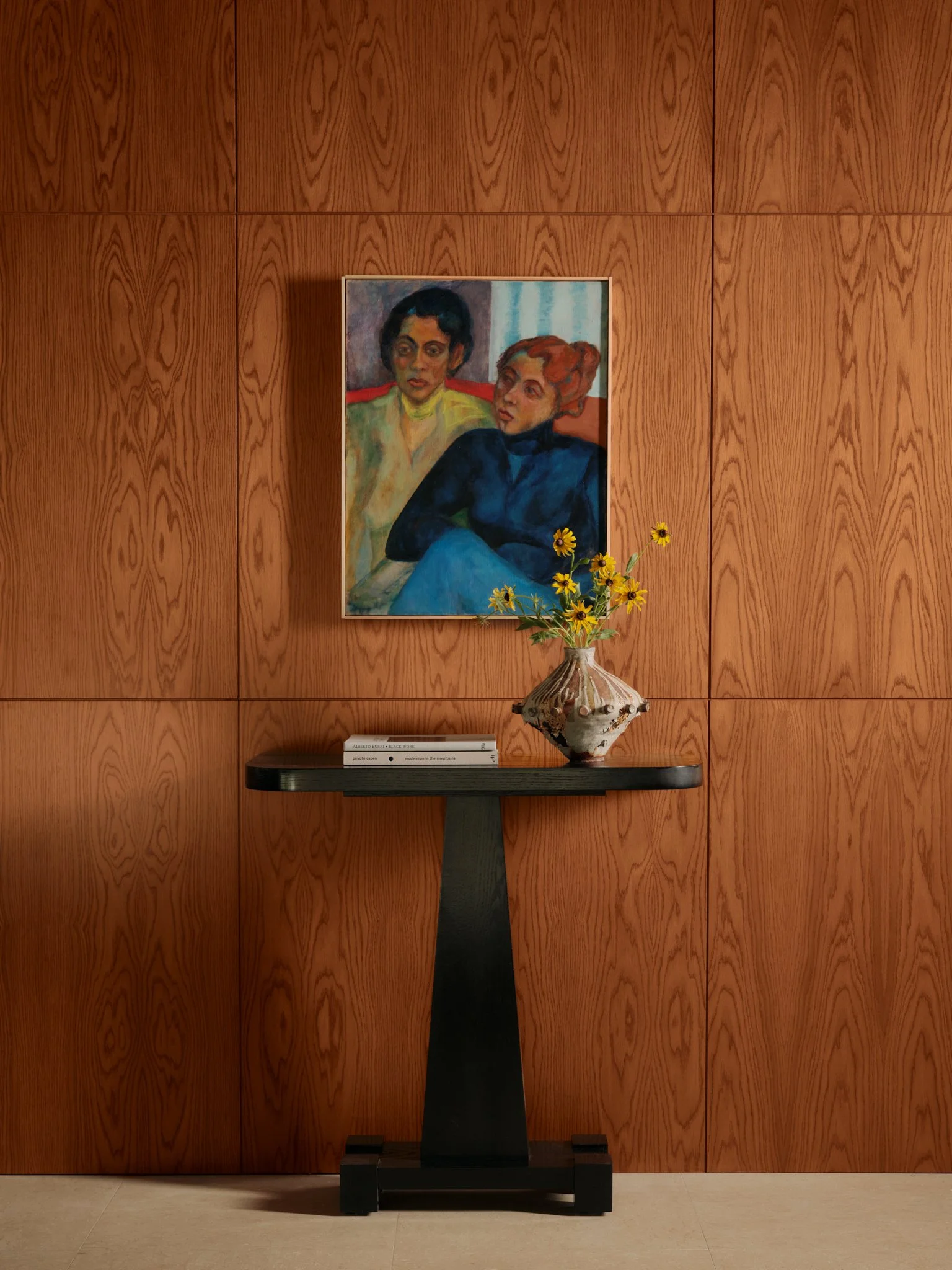 A wooden wall with a framed painting of two women, a black woman with short hair in yellow and green, and a white woman with red hair in dark clothing, behind a black table with a white book and a vase with yellow flowers.