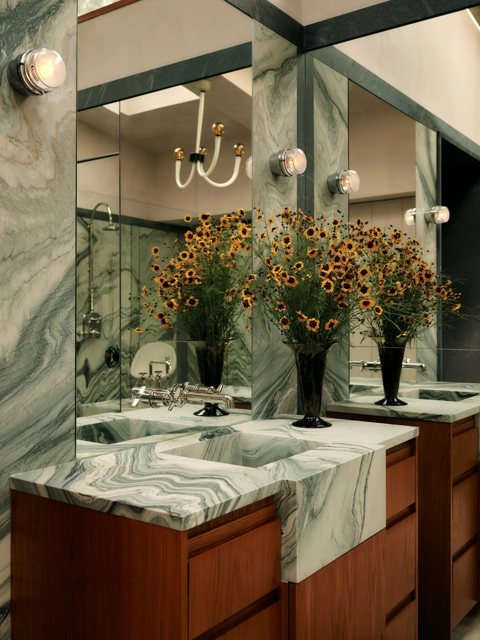 A marble bathroom vanity with two sinks, mounted mirrors, a white chandelier reflected in the mirror, and a bouquet of yellow flowers in a dark vase.