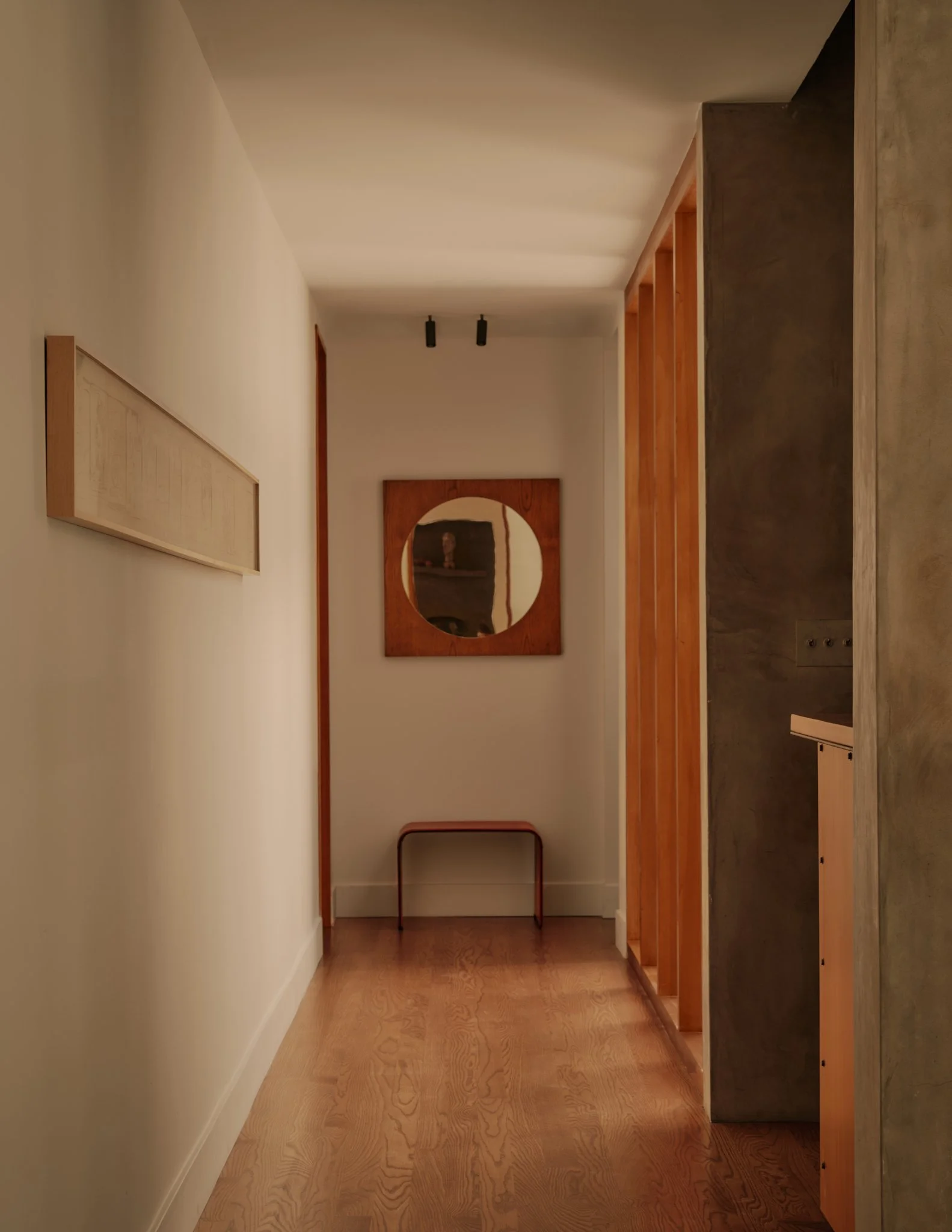 A hallway with wood flooring, beige walls, a framed sketch on the left wall, and a mirror with a round cutout on a wooden panel on the back wall. Black spotlights are mounted on the ceiling, and a small metal table is placed against the back wall.