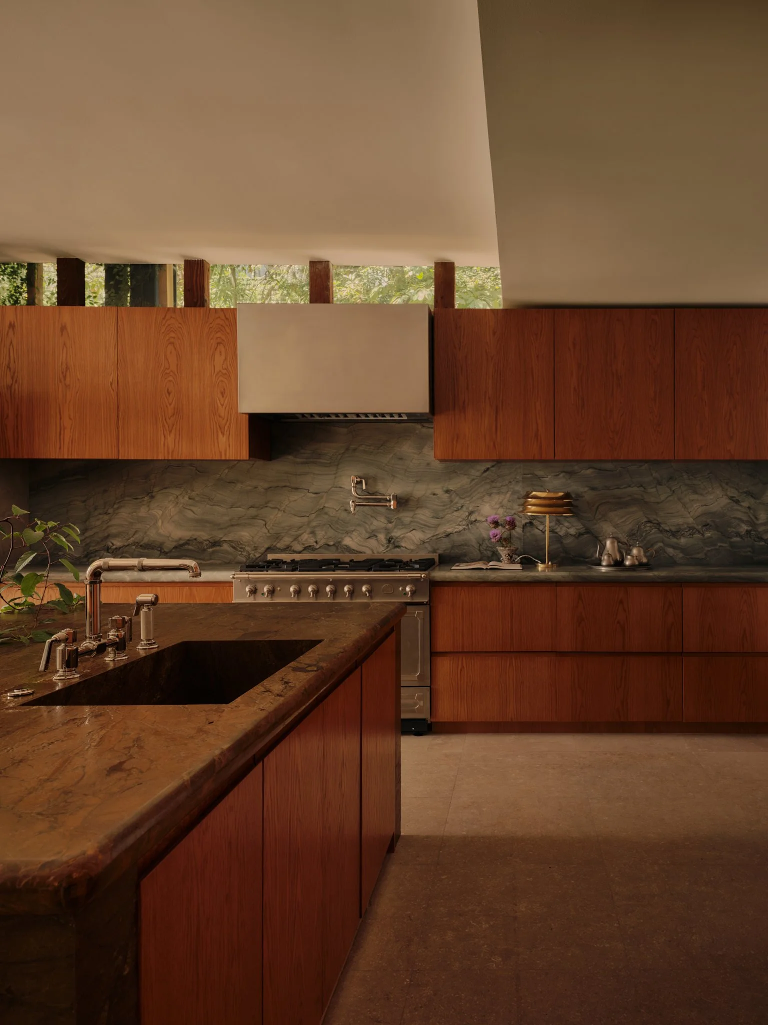 A modern kitchen with wooden cabinets, a marble backsplash, and a black sink on a brown marble countertop. The kitchen features a stove and a decorative lamp with purple flowers nearby.