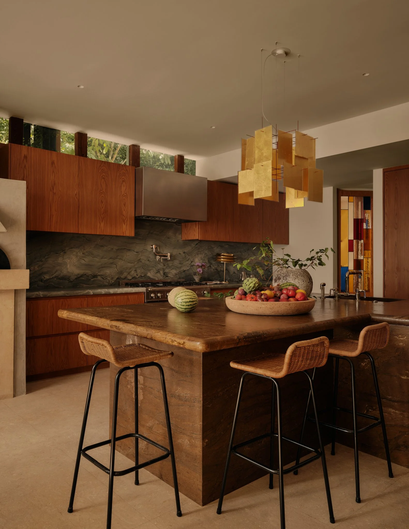 Modern kitchen with wooden cabinets, a black countertop, and a marble island. Hanging from the ceiling is a chandelier featuring gold and brown geometric panels. There are fruits and vegetables on the island and in a bowl. Wooden chairs with black legs are positioned around the island.