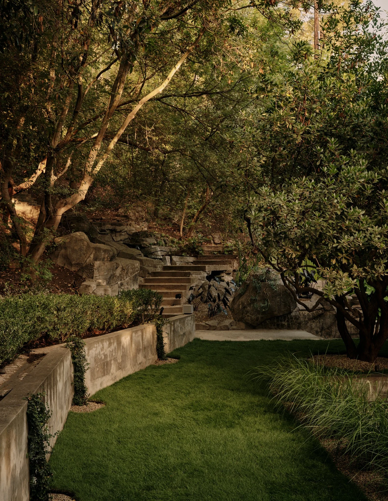 A landscaped backyard with stone steps, lush green grass, bushes, trees, and rocks, under a tree canopy.
