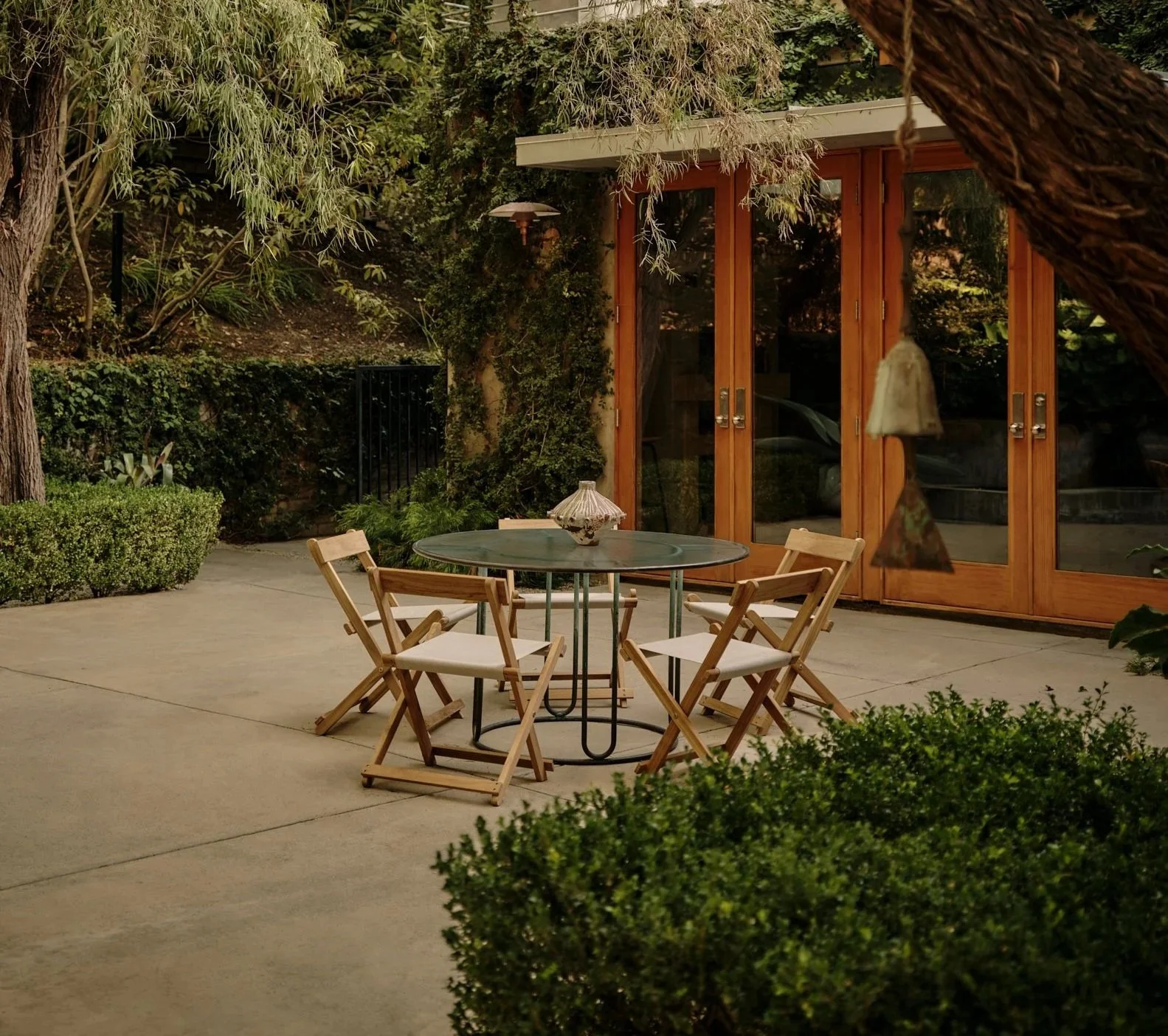 An outdoor patio with a round glass table, four wooden chairs with white cushions, a decorative lamp on the table, and a background of lush greenery and wooden-framed glass doors.