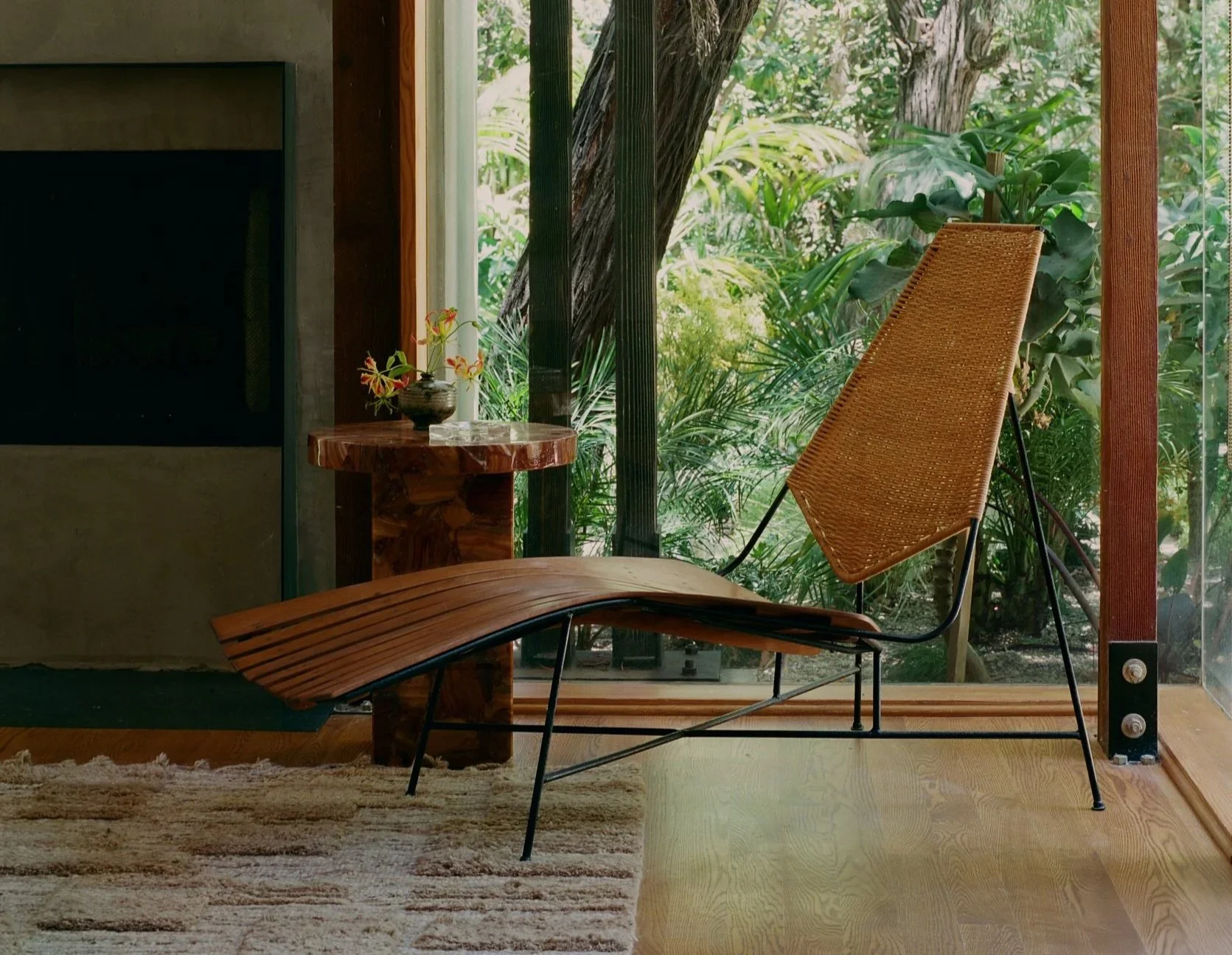 Living room with large window showing greenery outside, featuring a modern wicker lounge chair, a small round wooden side table with a plant, and a beige rug on light wooden flooring.