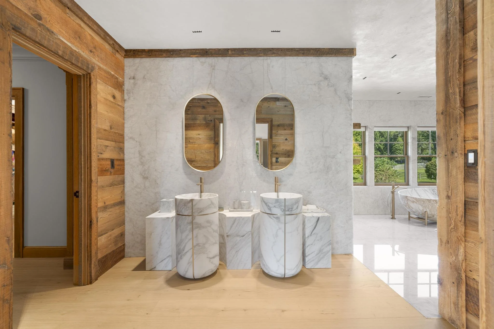 Modern bathroom with two marble pedestal sinks, two oval mirrors, large windows, wood trim, and natural light.