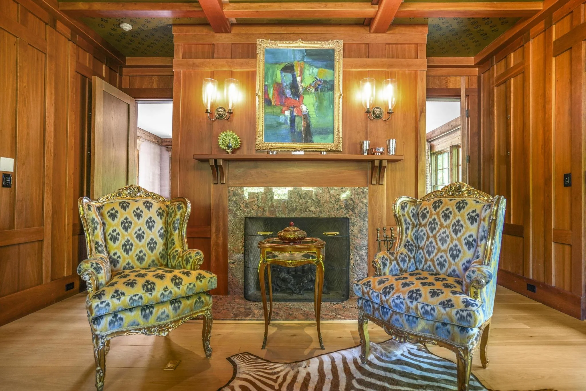 Two ornate yellow and black patterned armchairs with carved gold frames facing each other in front of a fireplace with a marble surround. Above the fireplace hangs an abstract colorful painting in a gold frame. There are two wall-mounted candle-style lights on either side of the painting, and decorative items on the mantel. The room has warm wooden paneled walls and ceiling, with a zebra-patterned rug on the floor in front of the fireplace.