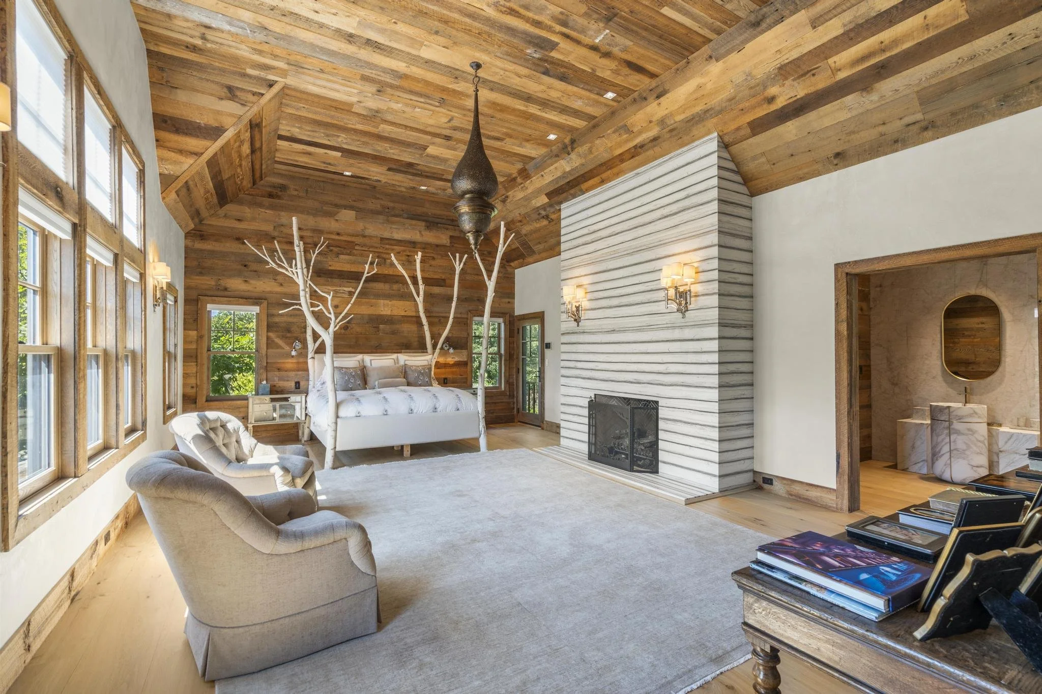 A spacious bedroom with a vaulted wooden ceiling and large windows. The room features a white bed with pillows and a white tree-shaped decoration on the headboard. Comfortable armchairs and a fireplace are present, with a stack of magazines on a wood