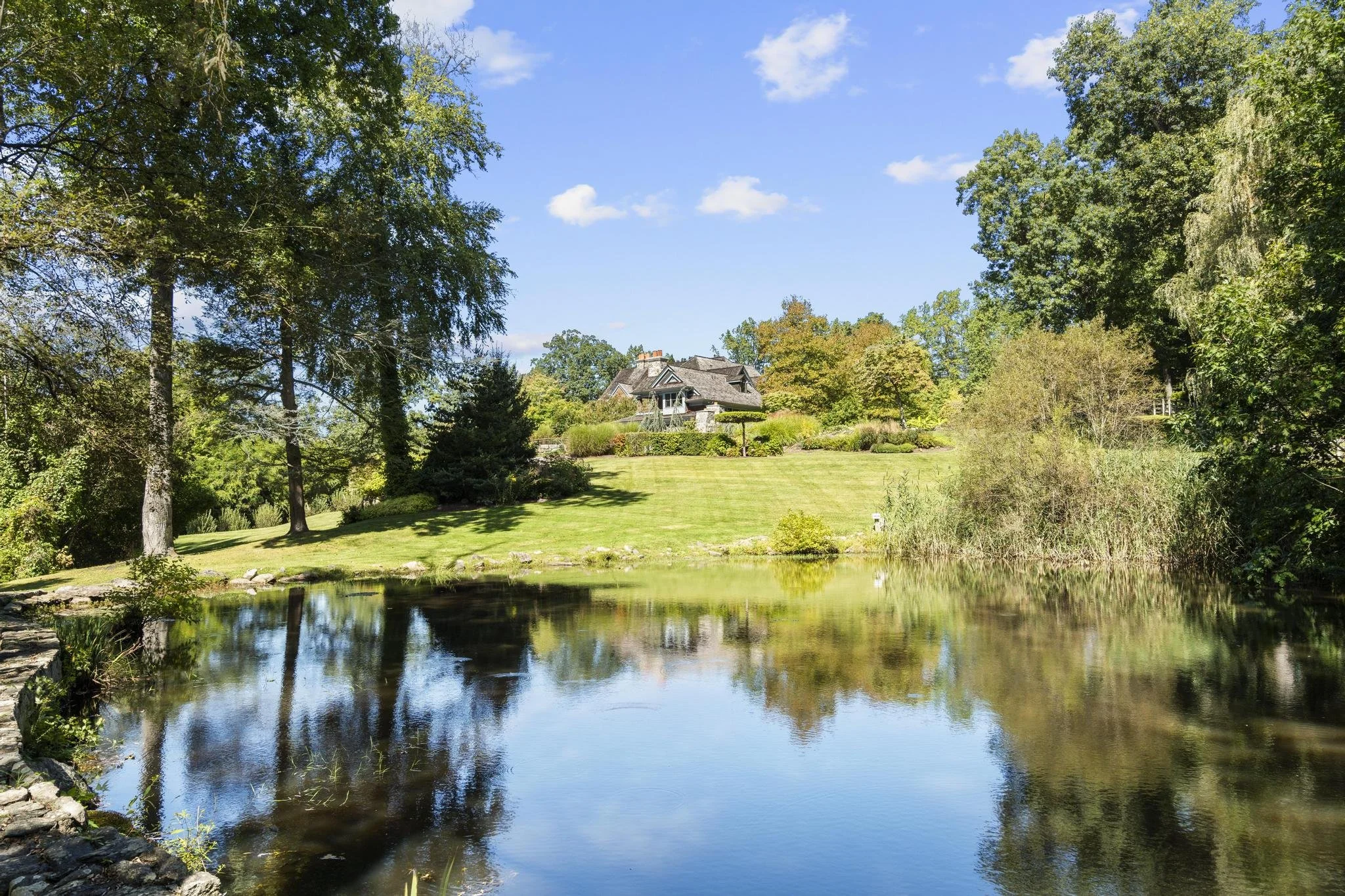 A house with a gray roof and chimney surrounded by green trees, overlooking a pond with reflections of the landscape and a clear blue sky with some clouds.