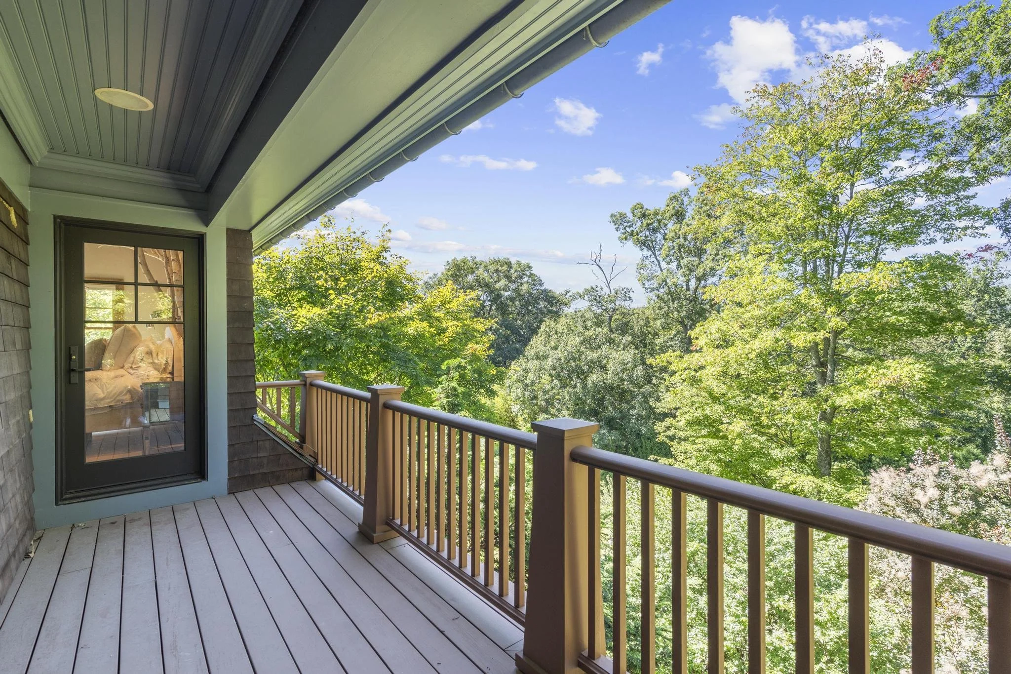 A balcony with a wooden railing overlooking a lush green tree-filled landscape under a blue sky with some clouds.