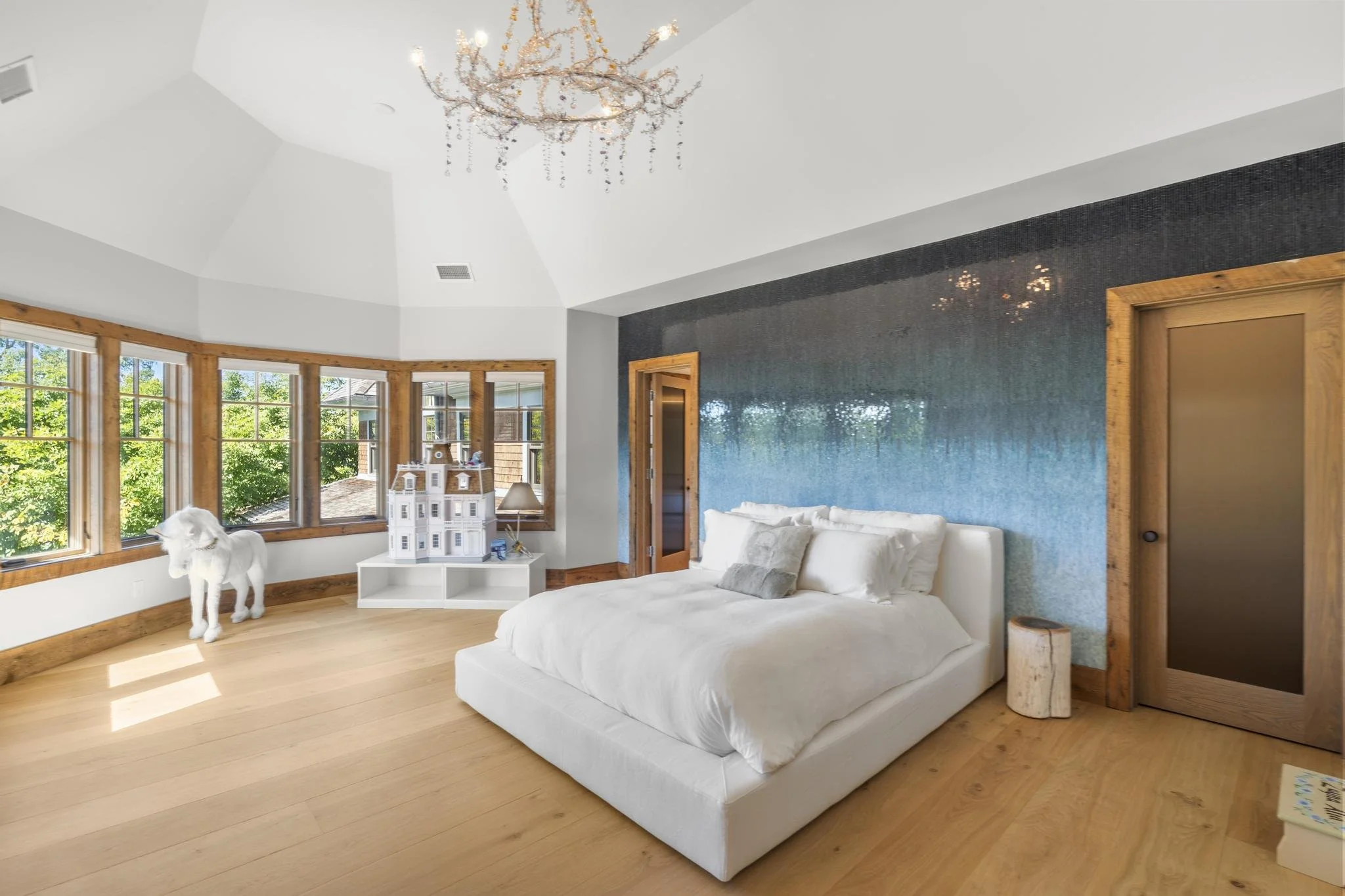 Bright bedroom with large windows, a white bed, a small white table with a dollhouse, a white horse sculpture, and a blue textured accent wall with a chandelier hanging from the ceiling.