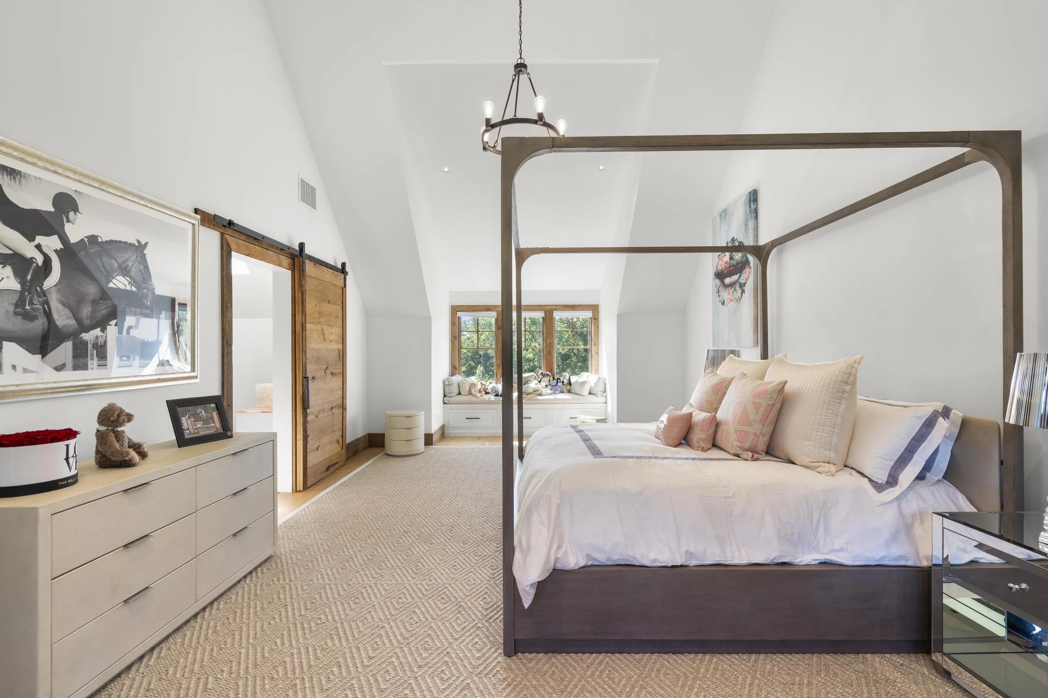 A bedroom with a bed featuring a wooden frame and pink pillows, a white dresser with picture frames and stuffed animals, a sliding barn door, and a window seat with cushions, decorated with artwork and a chandelier