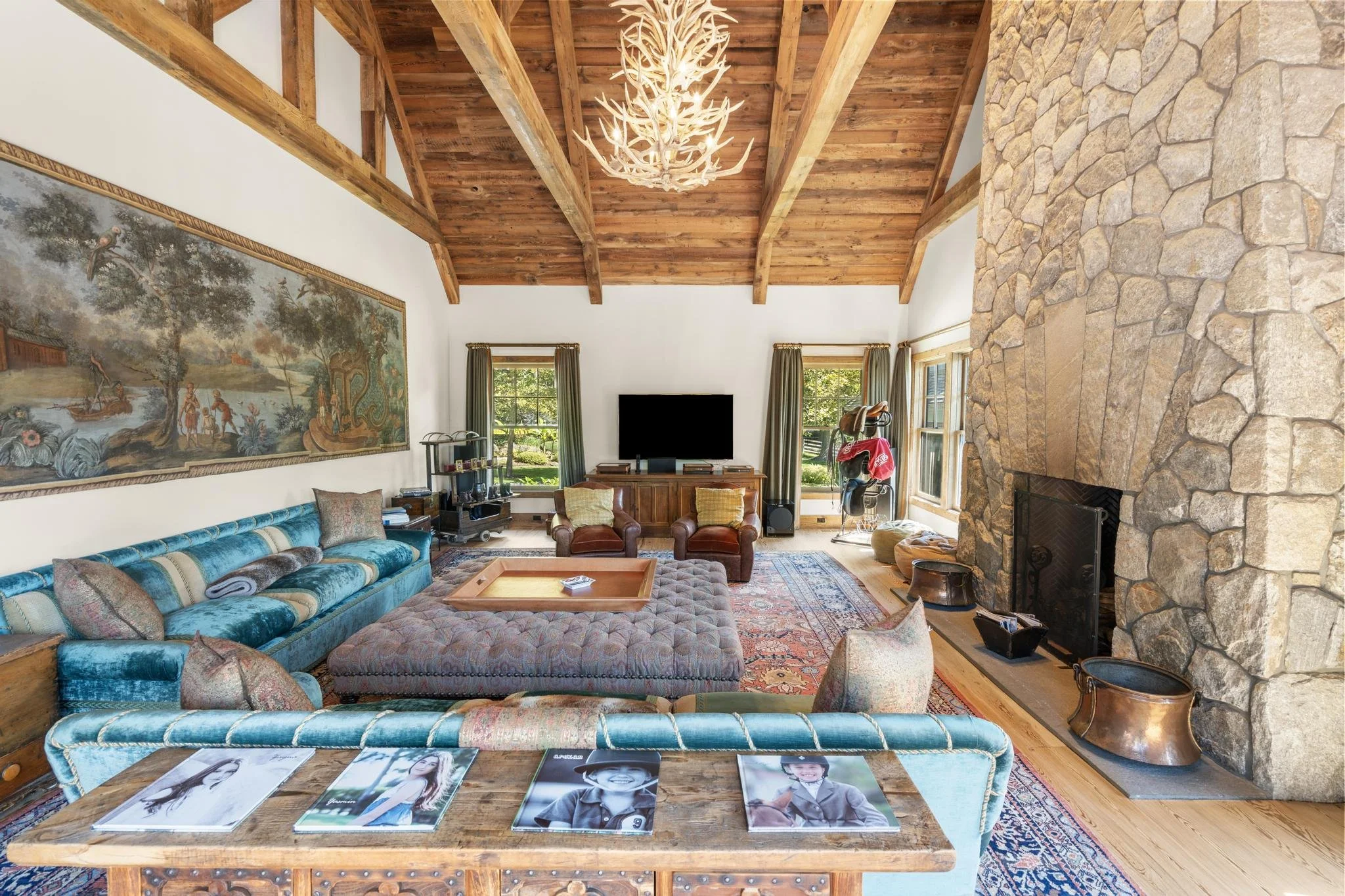 Living room with wooden vaulted ceiling, large stone fireplace, and multiple windows with green curtains, featuring furniture such as a blue velvet couch, plush ottomans, chairs, a wooden coffee table with magazines, and wall art of a pastoral landsc