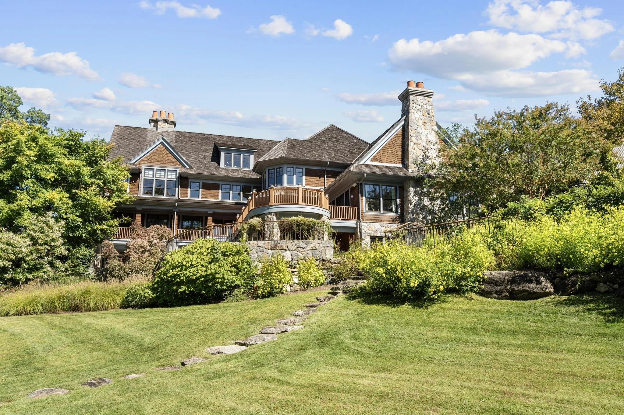 A large house with multiple stories, stone and wood exterior, surrounded by trees and a well-maintained lawn with stepping stones leading to the house, under a blue sky with scattered clouds.