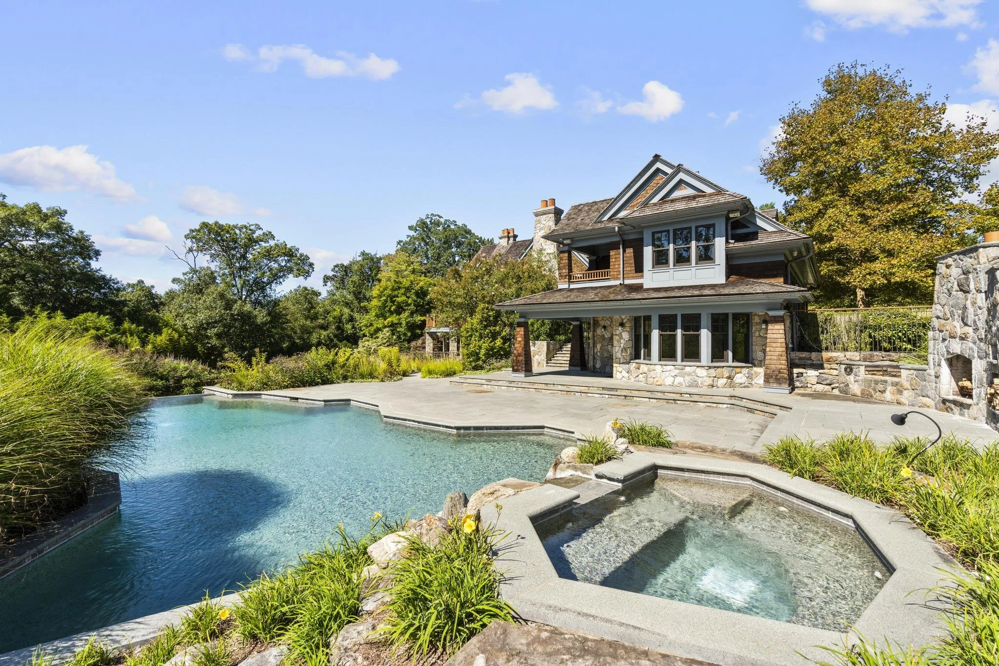 A house with a stone foundation, wooden siding, and multiple gabled roofs, situated next to a large swimming pool with surrounding greenery under a bright blue sky with scattered clouds.