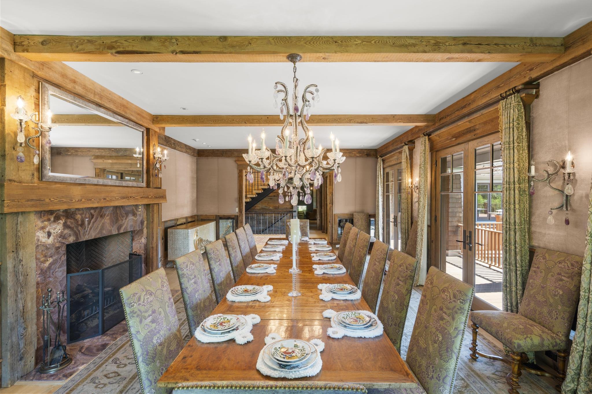 A dining room with a long wooden table set with plates and napkins, surrounded by patterned upholstered chairs. A large crystal chandelier hangs above the table, and the room features wood beams, a fireplace with a stone mantel, and large windows wit