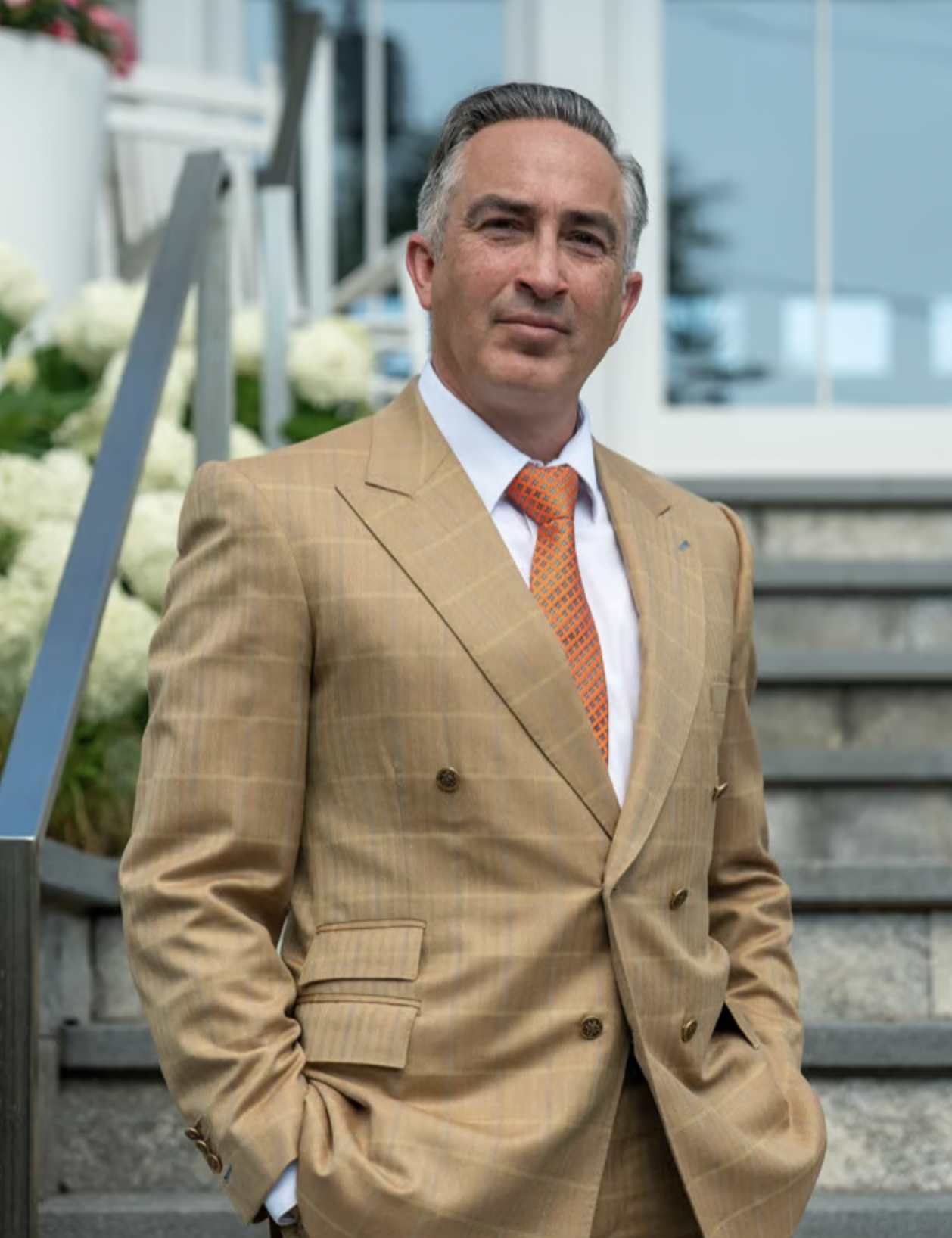 A middle-aged man with gray hair, dressed in a tan plaid suit and orange patterned tie, standing outdoors on a staircase with white flowers and a building in the background.