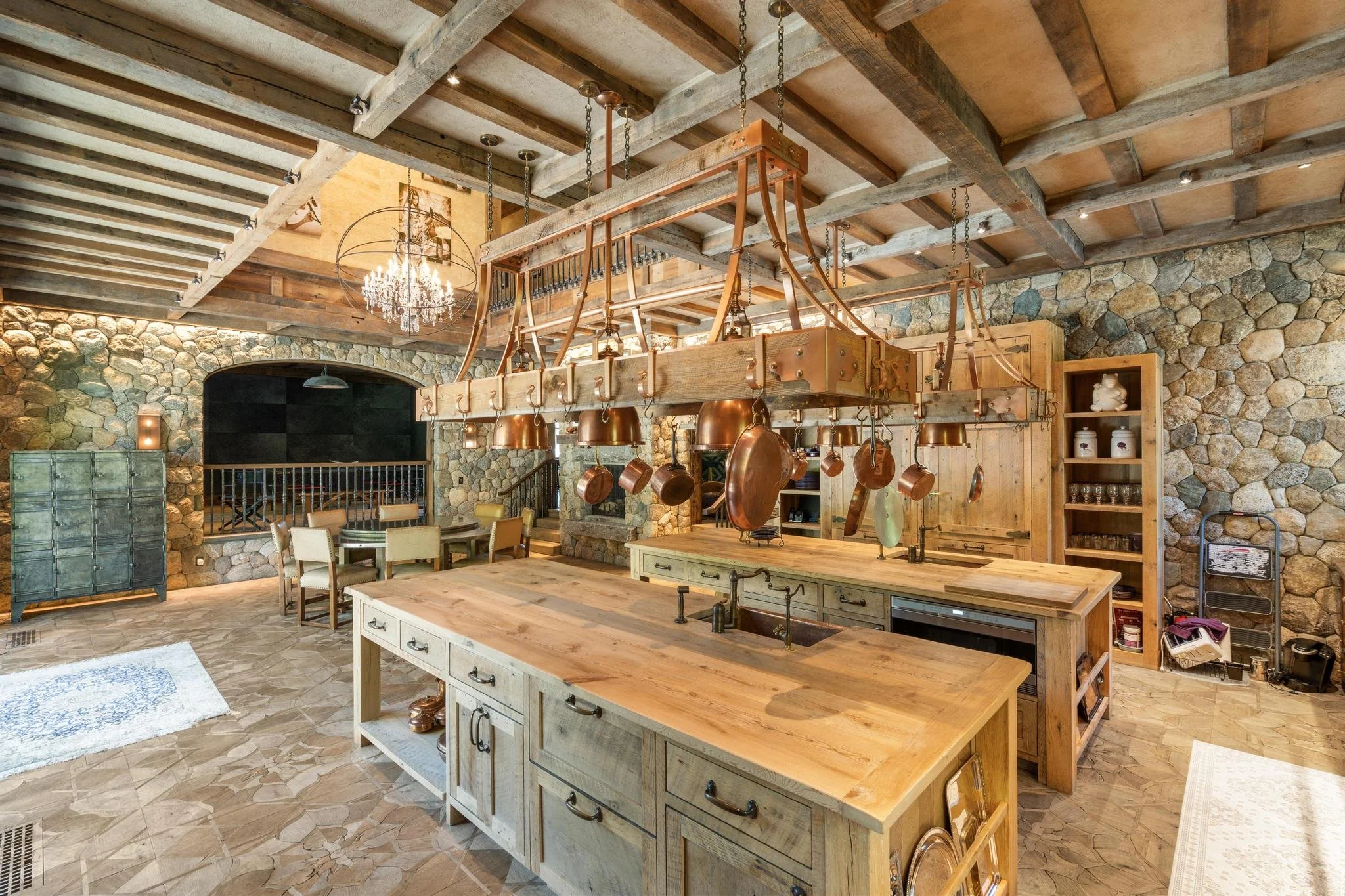 A rustic kitchen with a stone accent wall, wooden beams, and a chandelier hanging from the ceiling. A large wooden island with a sink is in the center, surrounded by chairs and kitchen utensils.