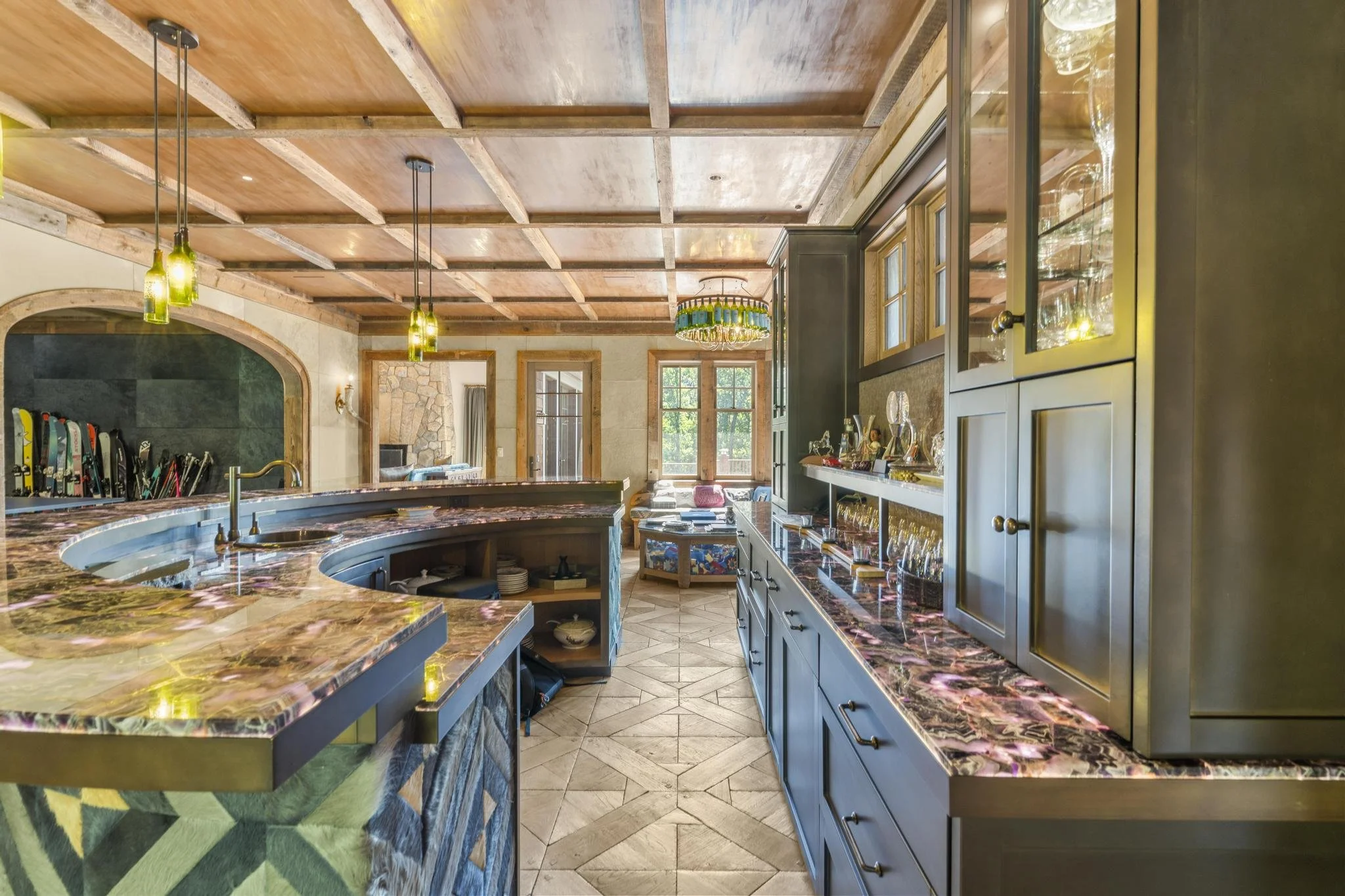 A spacious kitchen with wooden ceiling beams, pendant lights, large windows, and a granite countertop bar. Blue cabinetry with glass doors displays glassware and decorative items. A herringbone-patterned tile floor and a stone fireplace are visible in the background.