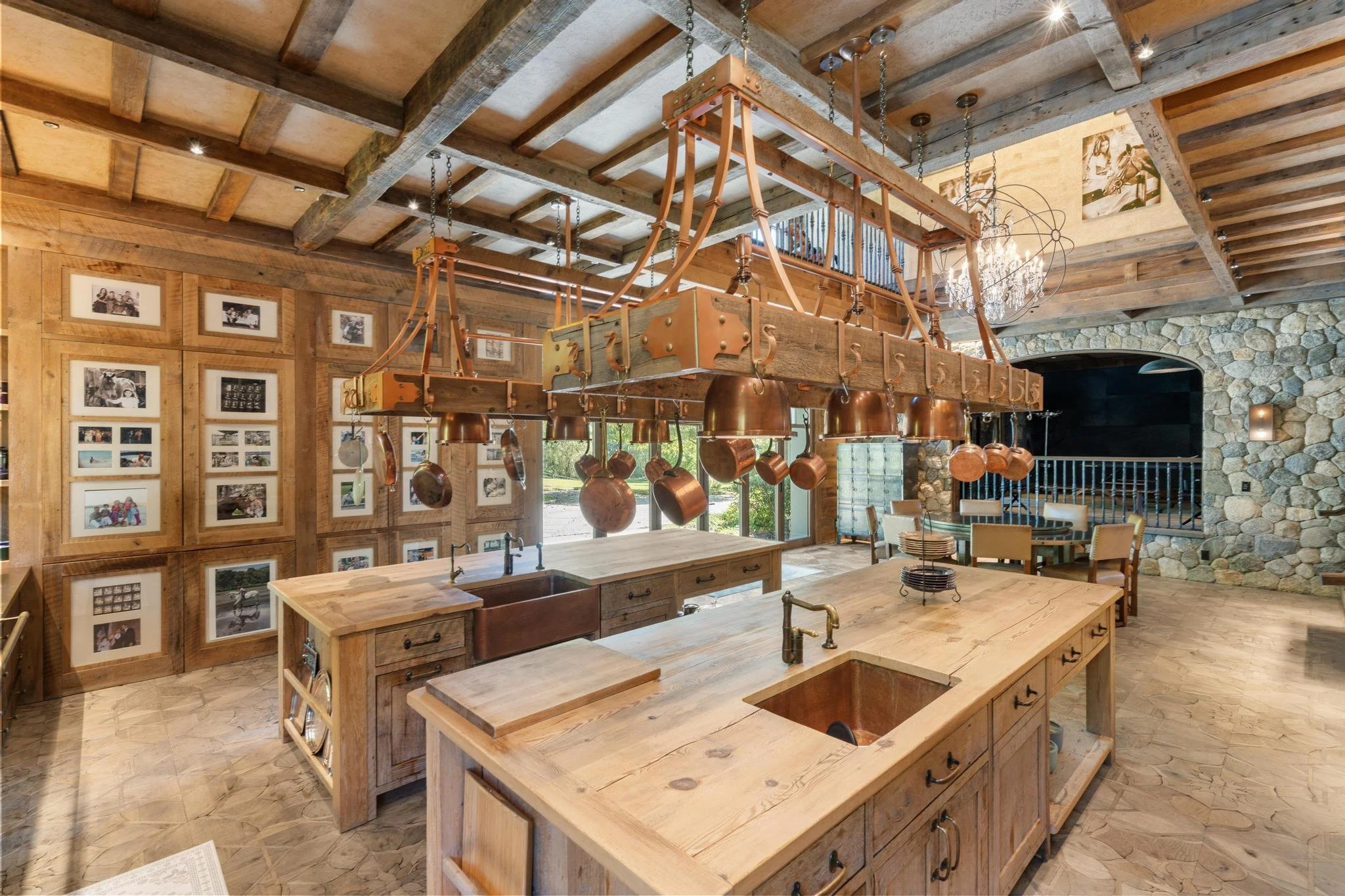 A rustic kitchen with wooden beams on the ceiling, a chandelier, and a large kitchen island with multiple copper pots hanging from a custom wood pot rack. The walls are decorated with framed photos and a stone fireplace is visible in the background.