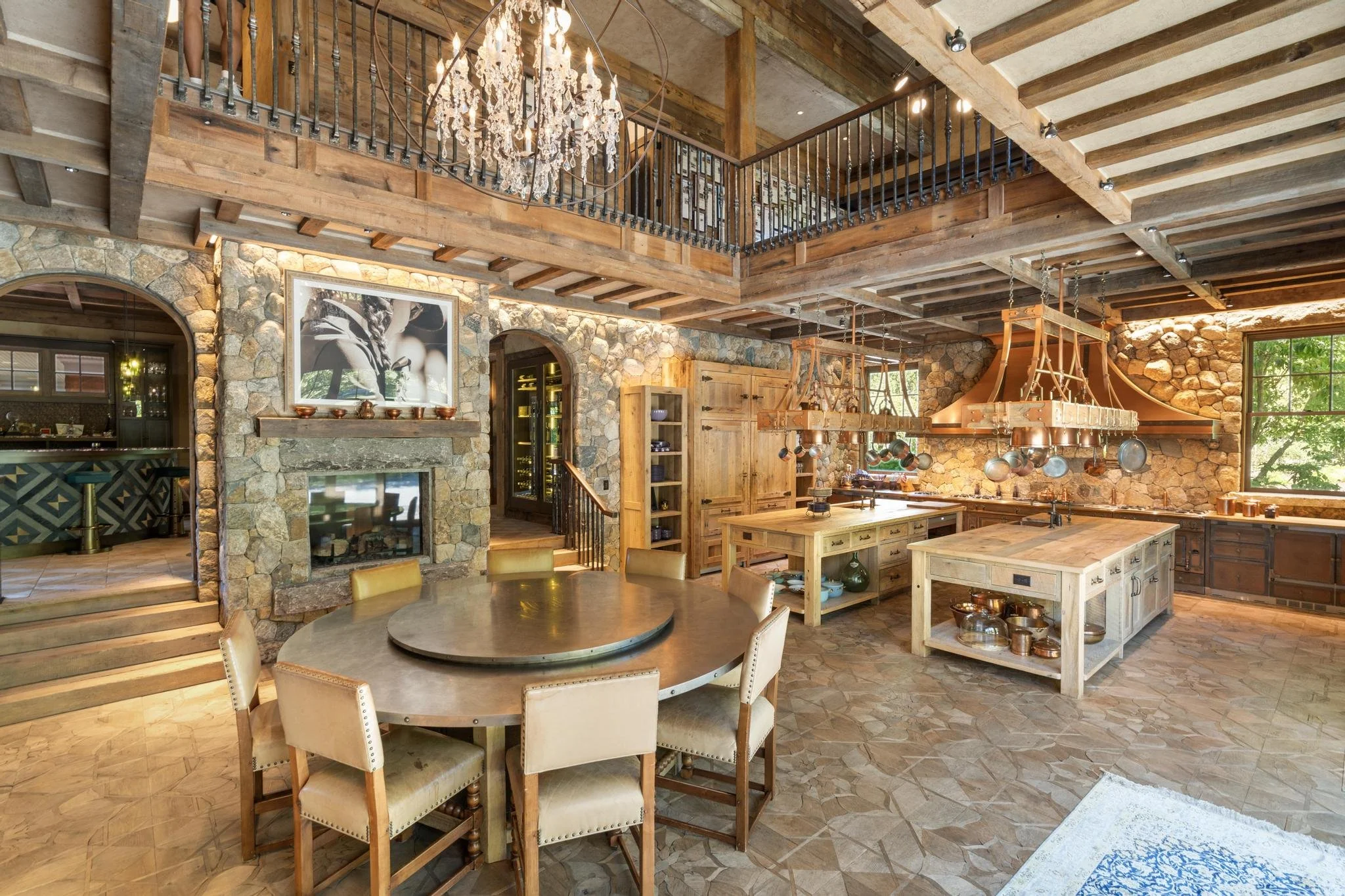 Interior view of a rustic kitchen with wood beams, stone walls, a chandelier, a ceiling with exposed wooden beams, a large round dining table with chairs, and a kitchen island with hanging pots.