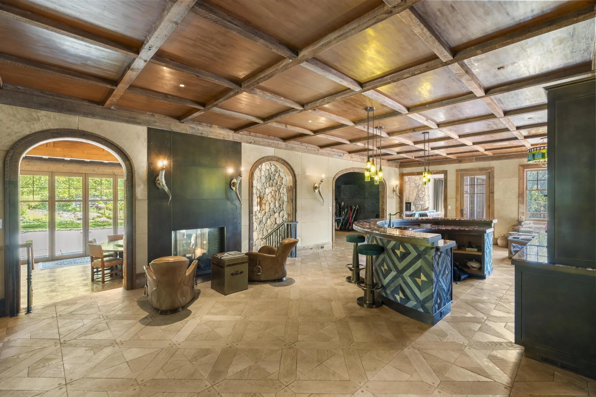 Interior view of a spacious home with wooden coffered ceiling, large windows with greenery outside, a stone fireplace flanked by wall sconces, a kitchen island with pendant lights, and various furniture.