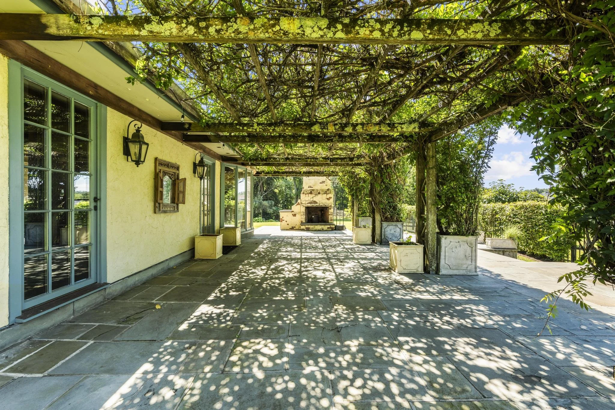 A patio area with a yellow house wall, green window frames, and black outdoor wall lights. Overhead, a wooden trellis with lush green foliage provides shade, casting intricate shadow patterns on the stone patio flooring. At the far end, there is a brick outdoor fireplace and potted plants line the perimeter.