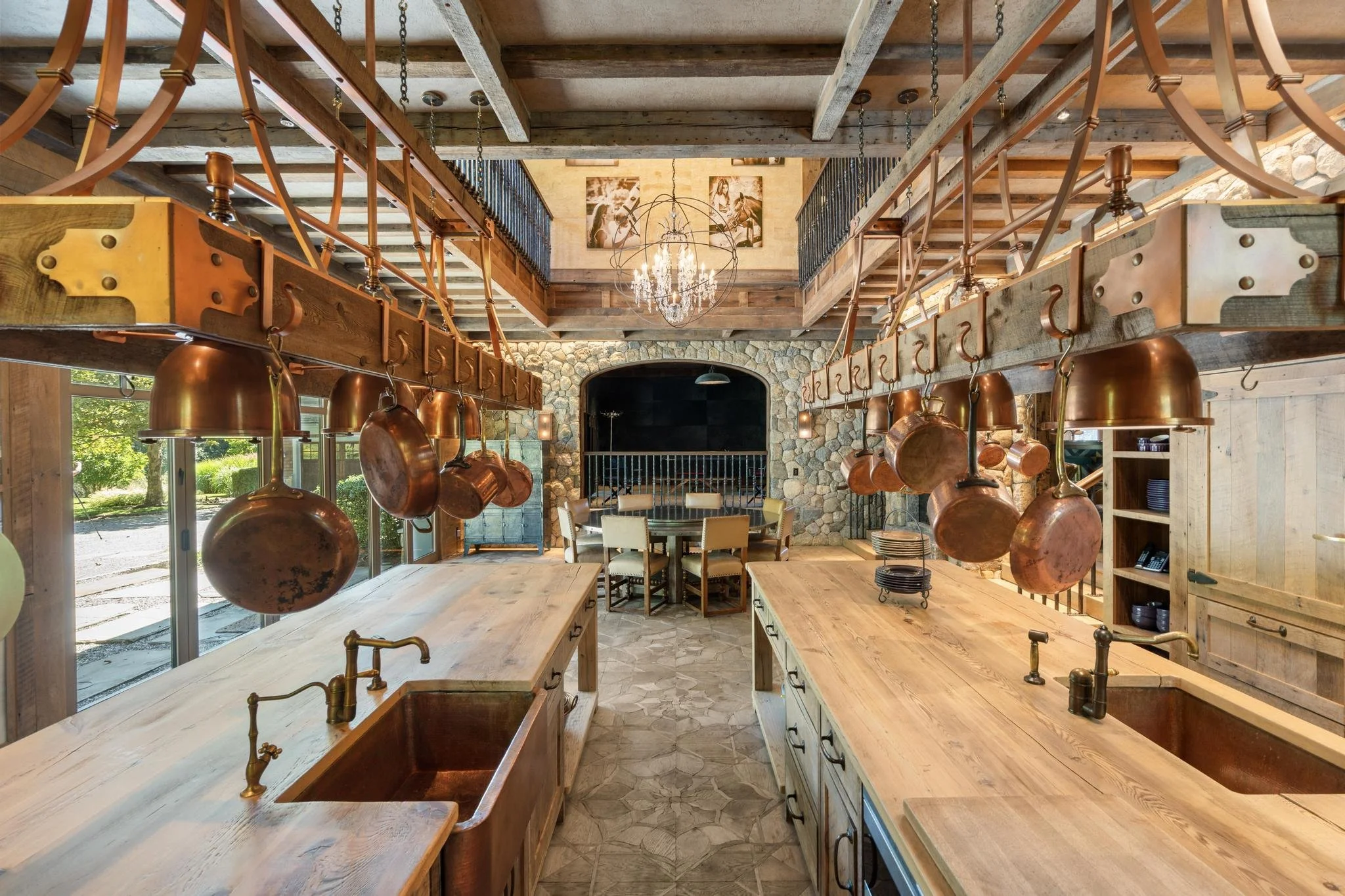 Interior of a rustic kitchen or dining area with wooden countertops and hanging copper pots. The room features a high ceiling with exposed wood beams, a stone accent wall with a fireplace, and large windows allowing natural light to illuminate the sp