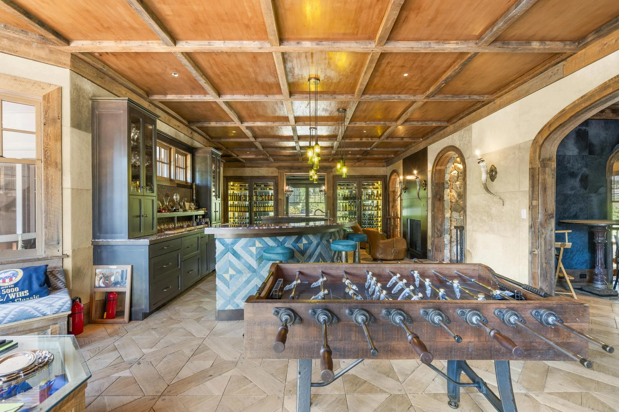 Interior of a rustic entertainment room with a wooden coffered ceiling, a bar with green cabinets, wine bottles, hanging pendant lights, a foosball table, and decorative elements.