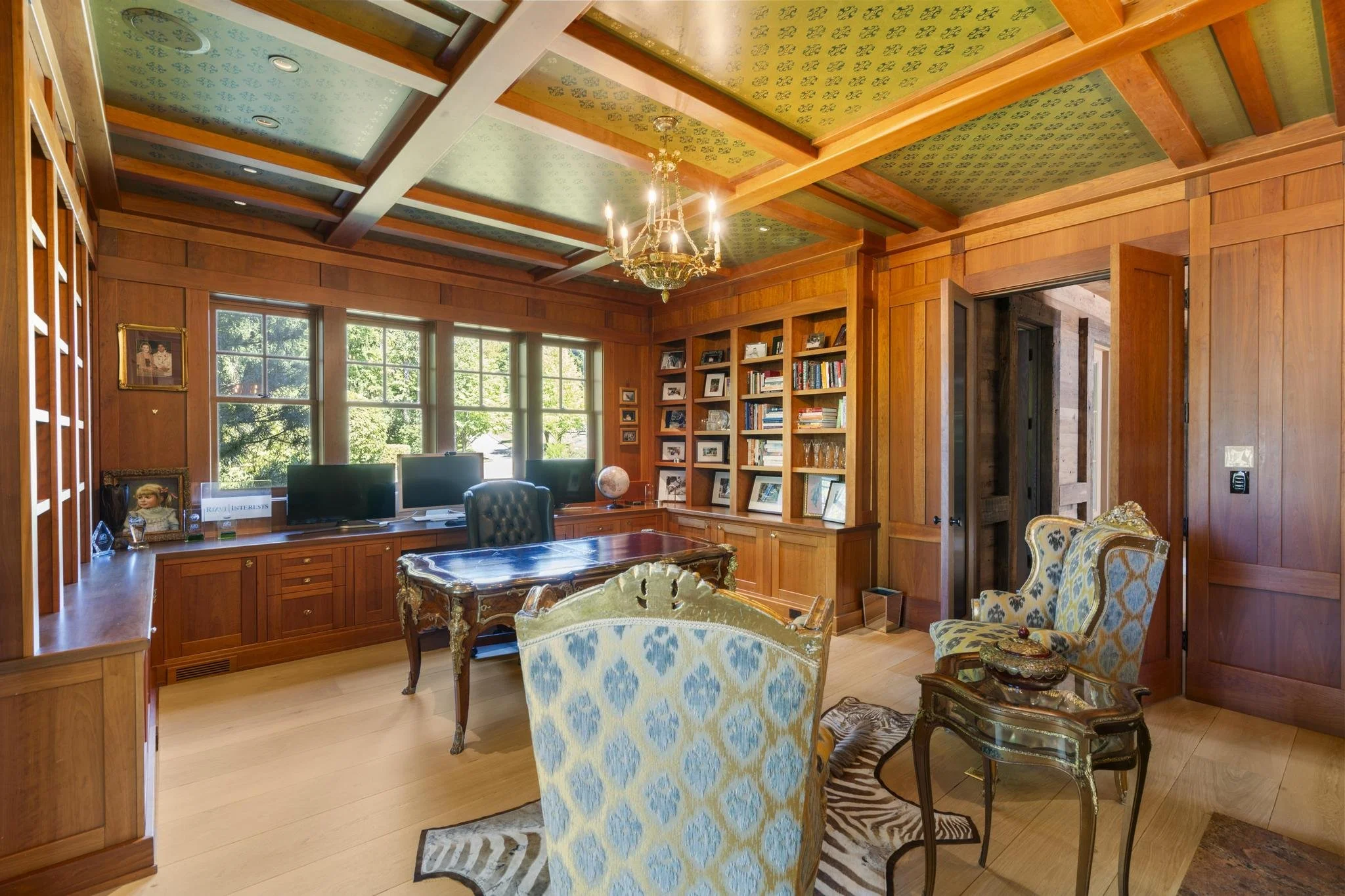 A cozy living room with wood-paneled walls and a wood-beamed ceiling, featuring large windows that let in natural light, a chandelier hanging from the ceiling, a patterned armchair, a glass table, and a built-in bookshelf with framed photos, books, a