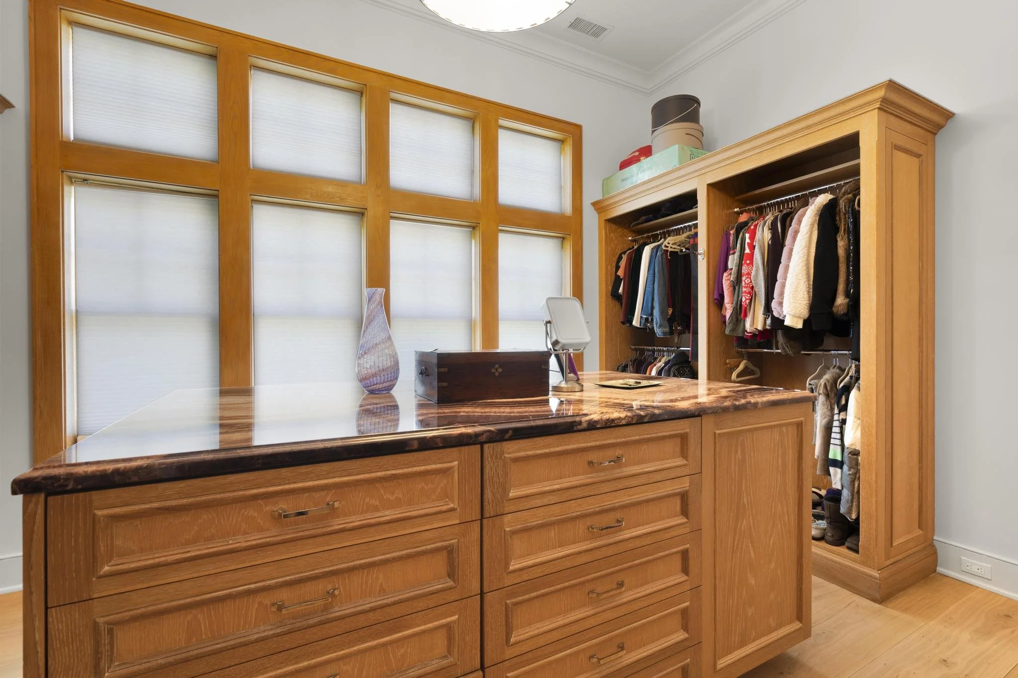 A wooden dresser with multiple drawers, topped with a decorative vase and a jewelry stand, situated next to a built-in wooden wardrobe with hanging clothes outside and boxes on top, in a well-lit room with white walls and windows with blinds.