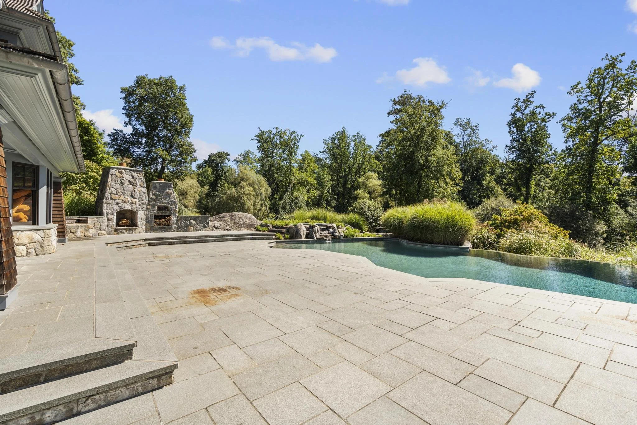 A backyard patio with an outdoor fireplace made of stone, a swimming pool, and lush green trees in the background under a blue sky with some clouds.