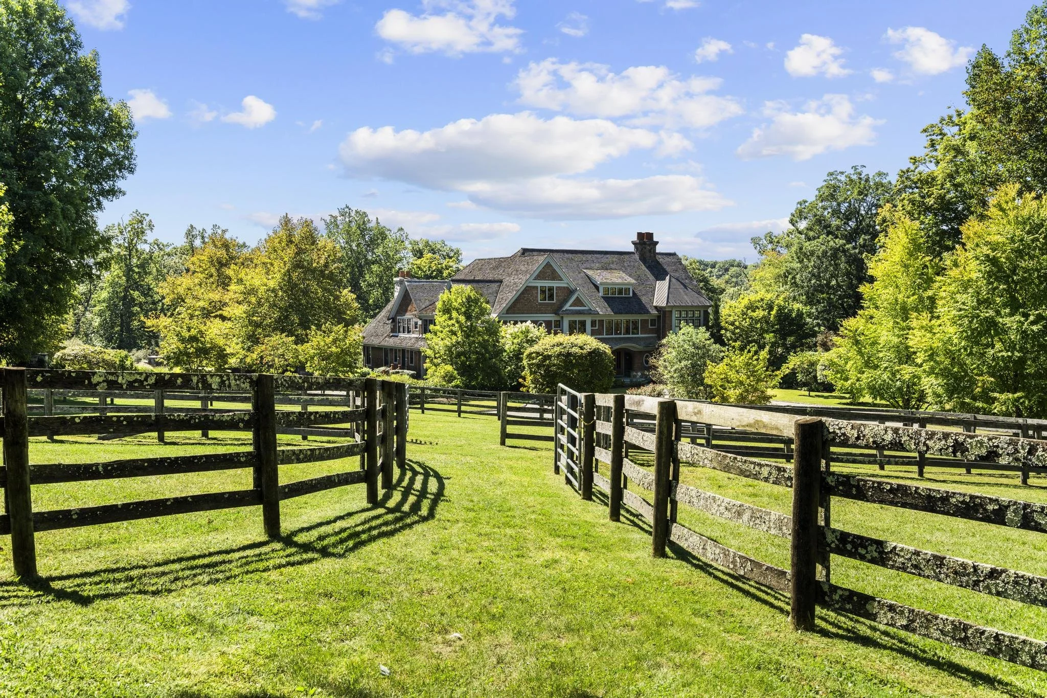 A large house with a dark roof and multiple windows is surrounded by green trees, under a blue sky with scattered clouds. A wooden fence encircles a grassy yard in the foreground.