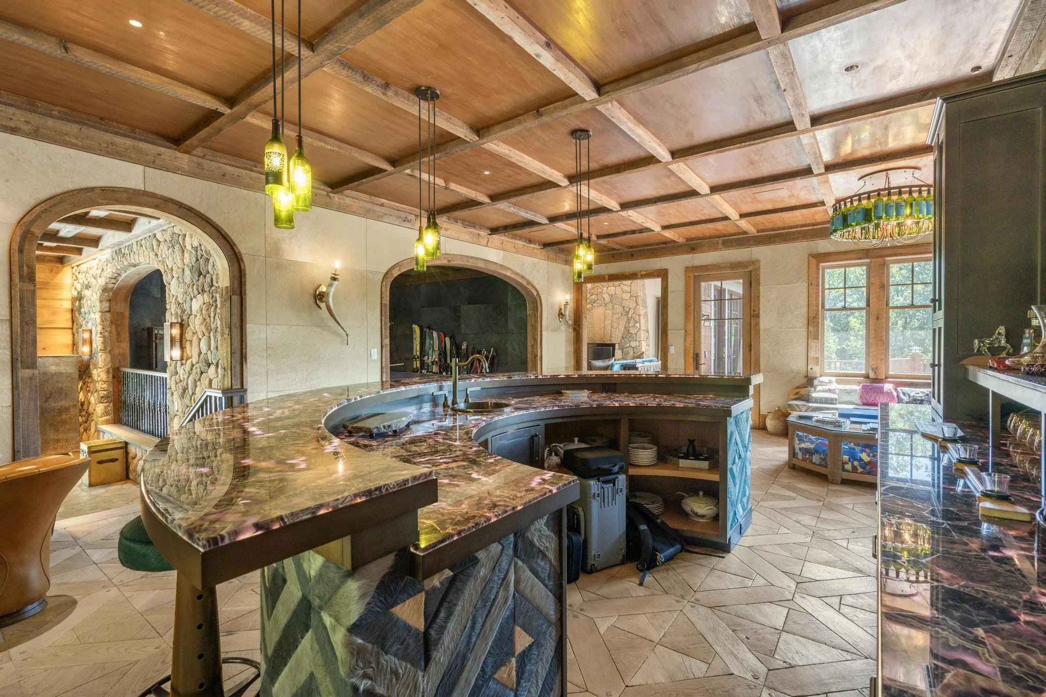 A cozy kitchen with a wooden coffered ceiling, hanging pendant lights, a marble-topped island, and large windows letting in natural light.