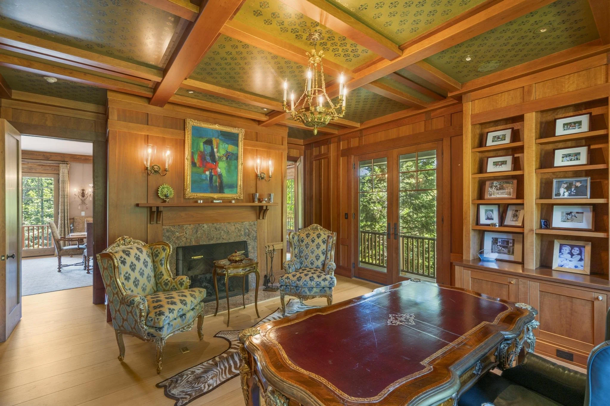 Living room with wooden walls and ceiling, featuring a chandelier, framed artwork, and a window with greenery outside. There are upholstered chairs, a fireplace, and a large wooden table.