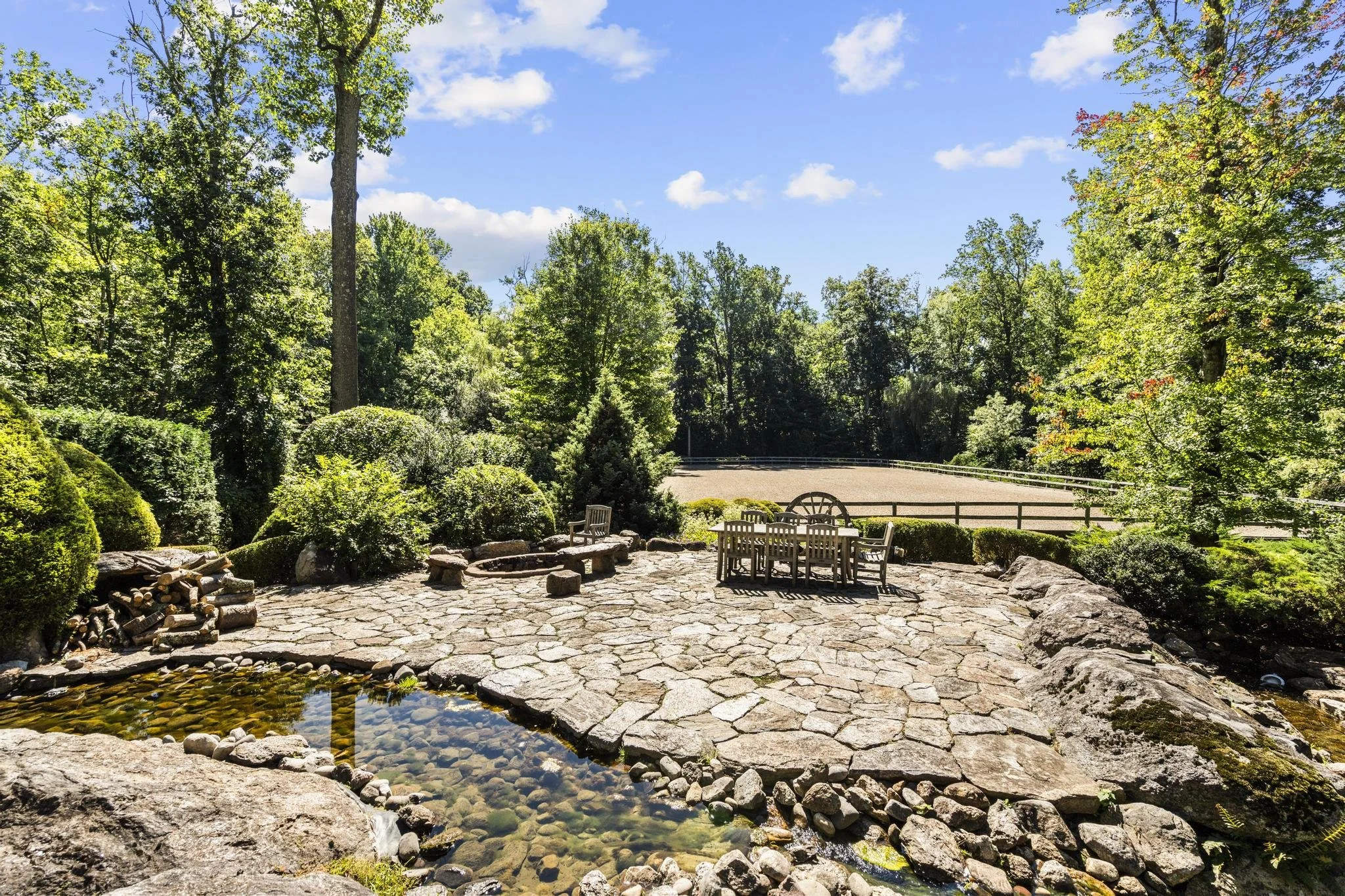 A scenic view of a park with lush green trees, a small pond with clear water, a stone patio area with outdoor furniture, and a wide dirt path surrounded by dense foliage under a partly cloudy sky.