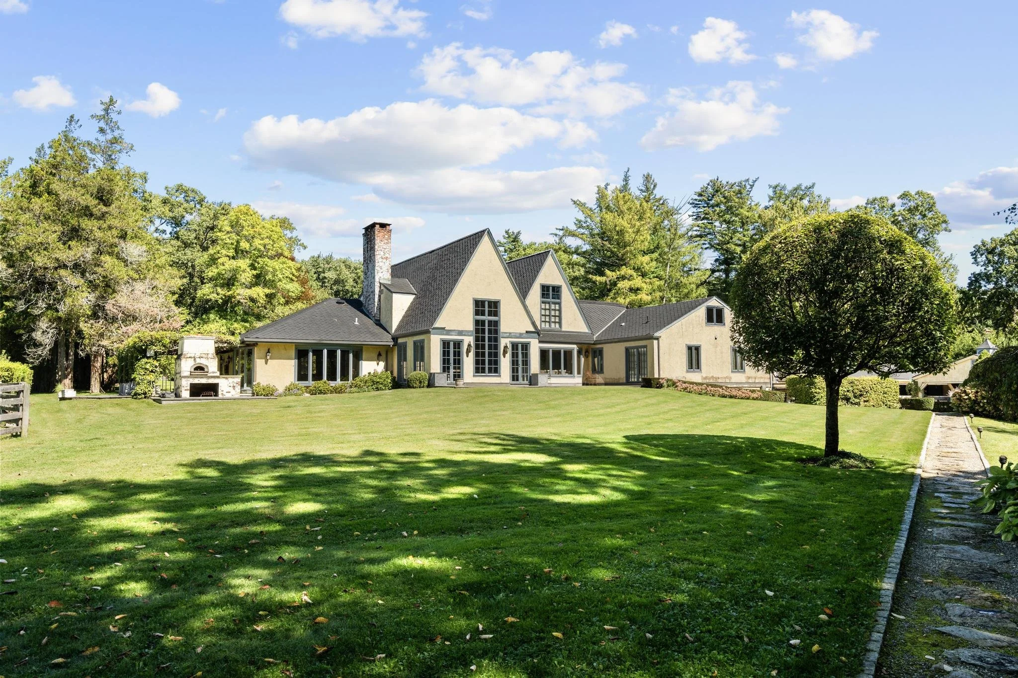 Large house with multiple gabled roofs, large windows, surrounded by a well-manicured lawn and trees, under a partly cloudy sky.