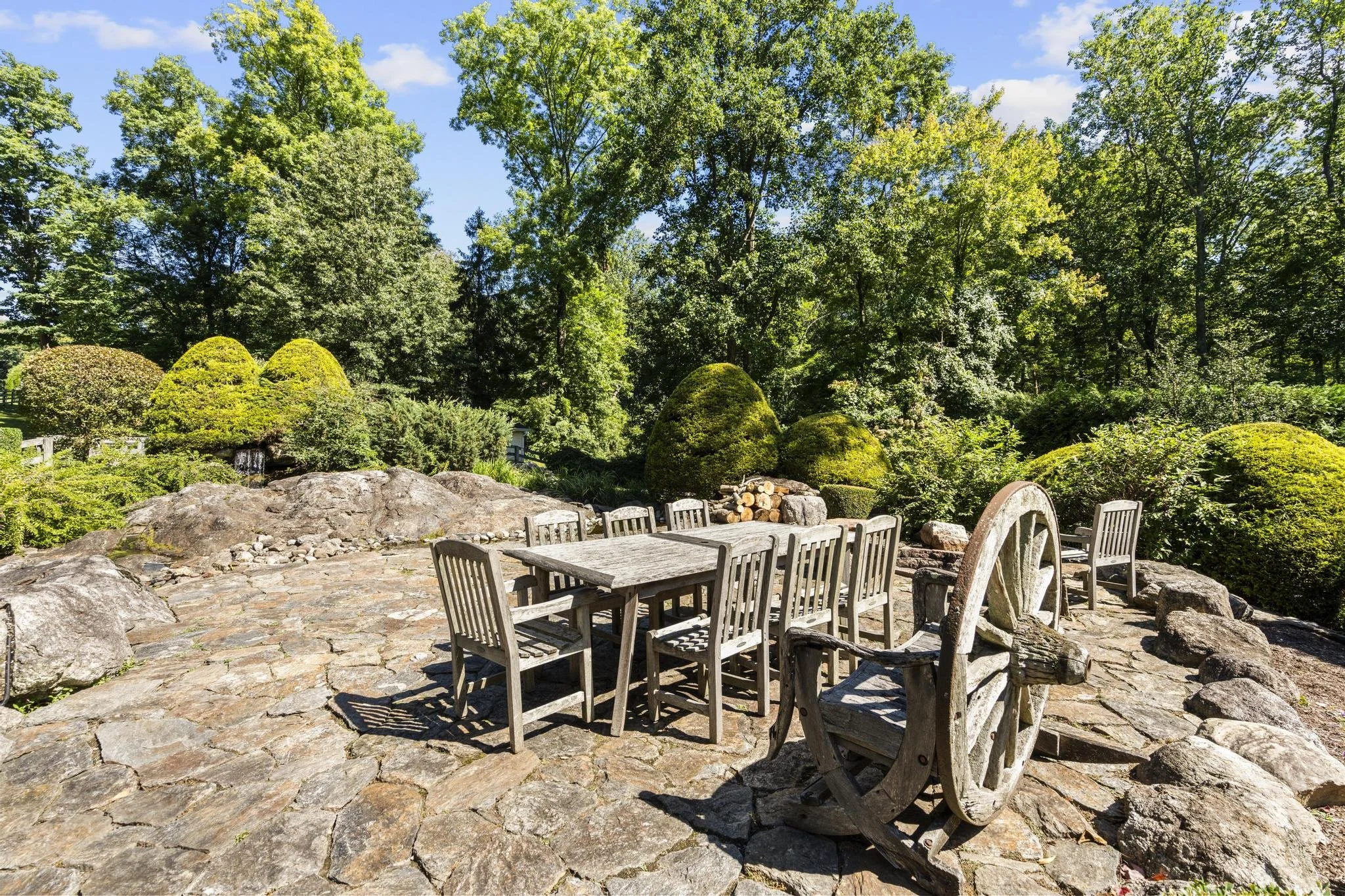 A landscaped outdoor area with a cobblestone patio, surrounded by lush green trees and shrubs, with a white wooden table and chairs, under a bright blue sky.