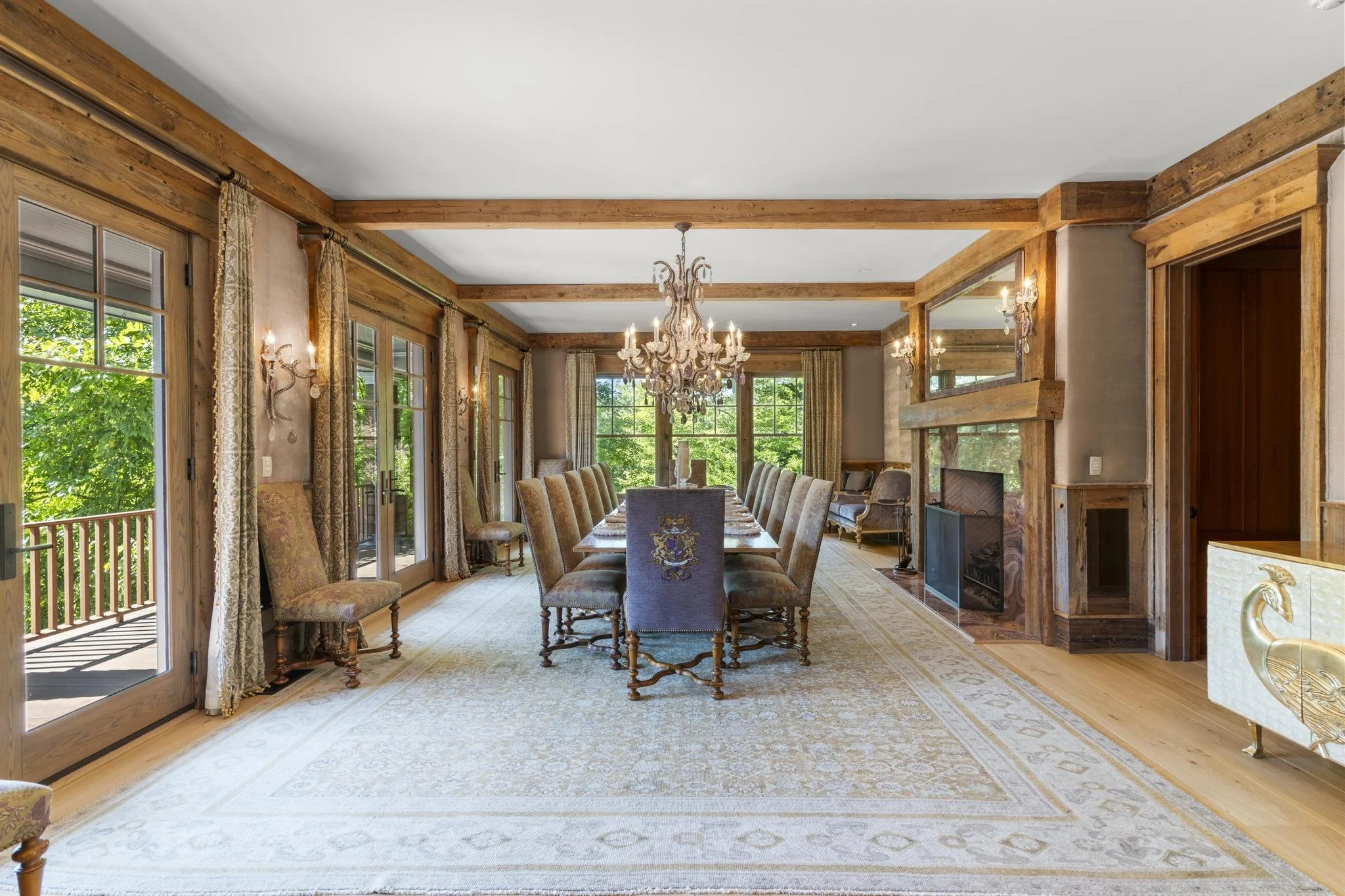 A dining room with wooden beams, a chandelier, a long dining table with upholstered chairs, large windows with curtains, and a view of outdoor greenery.