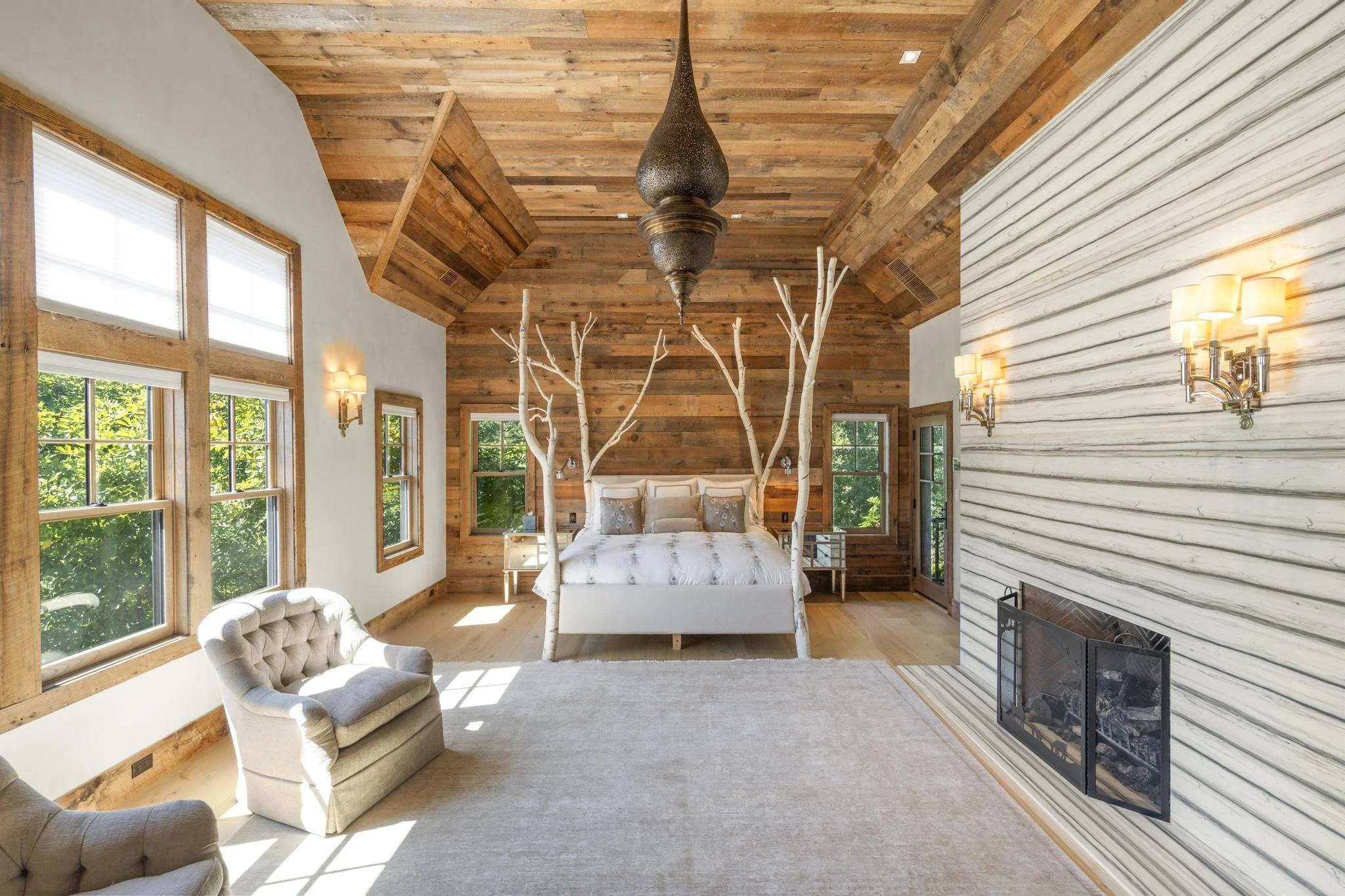 A bedroom with wooden paneled ceiling and walls, large windows with blinds, a bed with beige bedding, two white trees acting as headboard decor, a white tufted armchair near the window, a fireplace with a marble surround, and wall-mounted lamps.