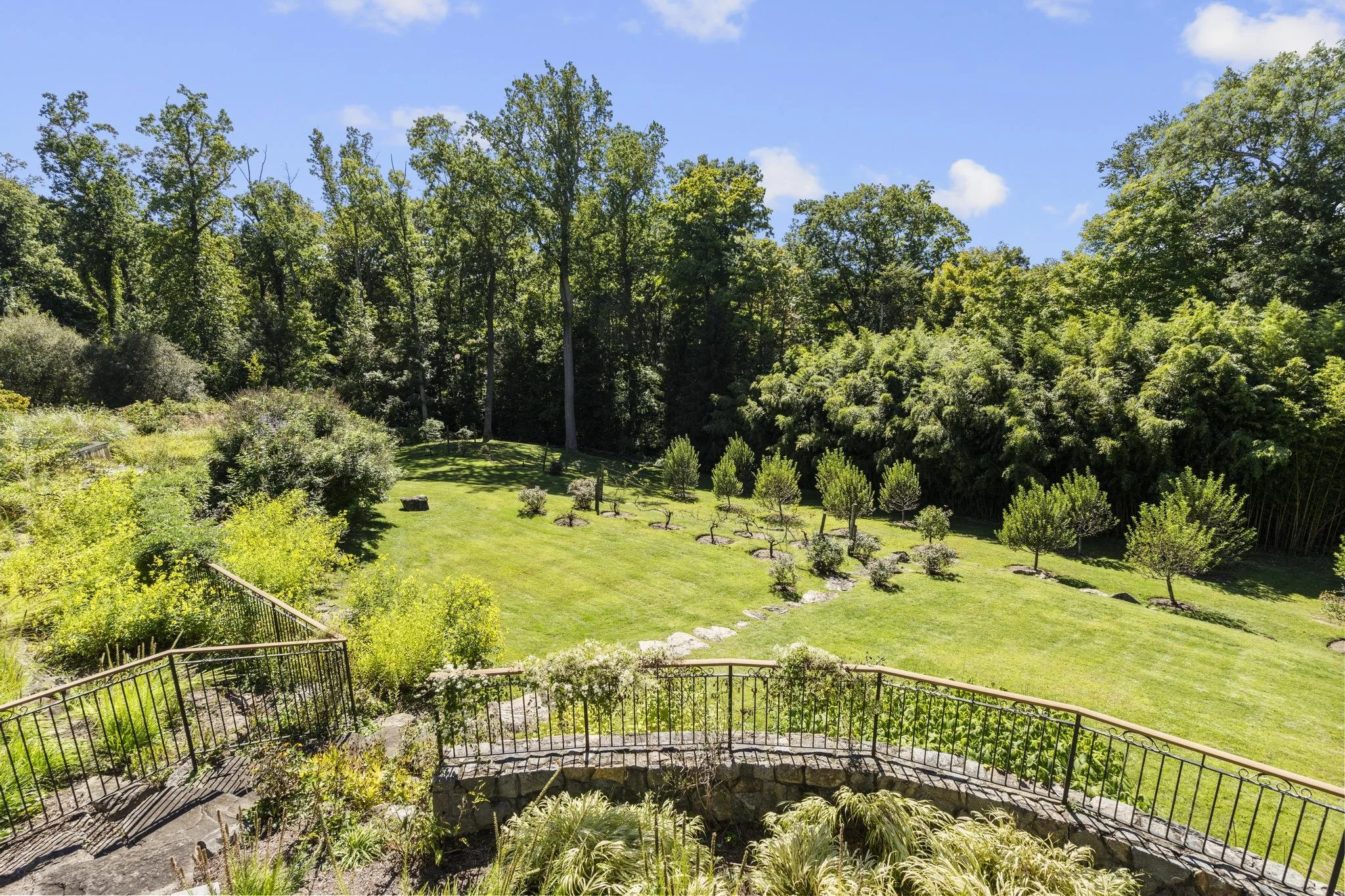 A garden with a curved stone pathway and black metal fence, lush green plants and trees, and a large grassy area with small trees, all under a blue sky with some clouds.