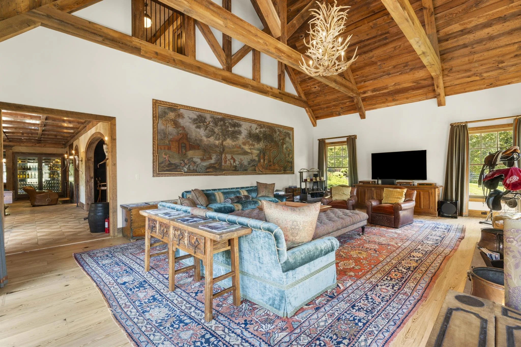 Quaker Lane Farm interior, great room. A spacious living room with a high, wooden vaulted ceiling and exposed beams. It features a large, ornate rug, vintage and modern furniture including a sofa, armchairs, and a leather ottoman.