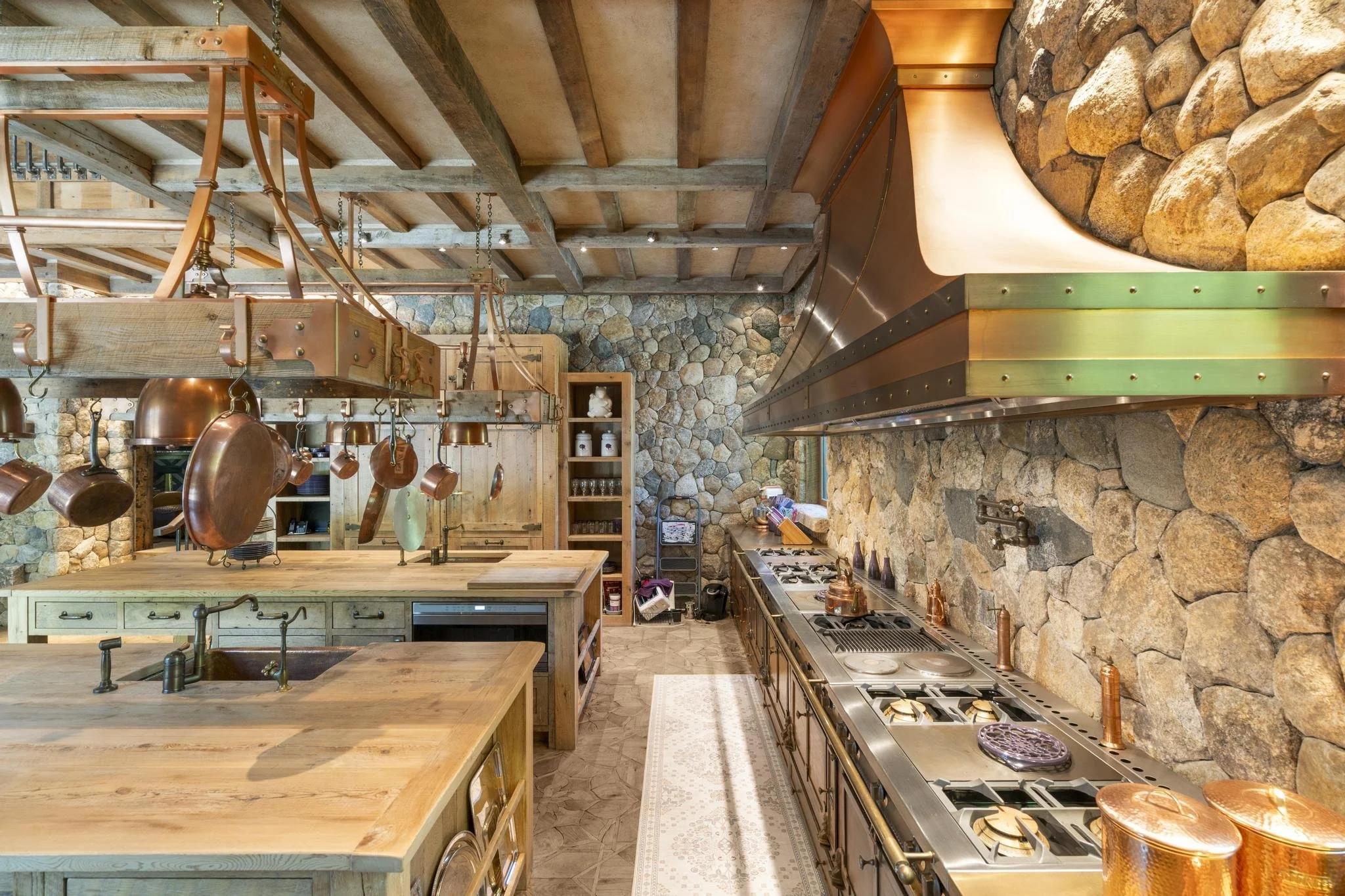 A rustic kitchen with stone walls, wooden beams on the ceiling, a large kitchen island with a wood countertop, hanging copper pots, a gas stove with copper knobs, and a wall-mounted range hood made of wood and metal.